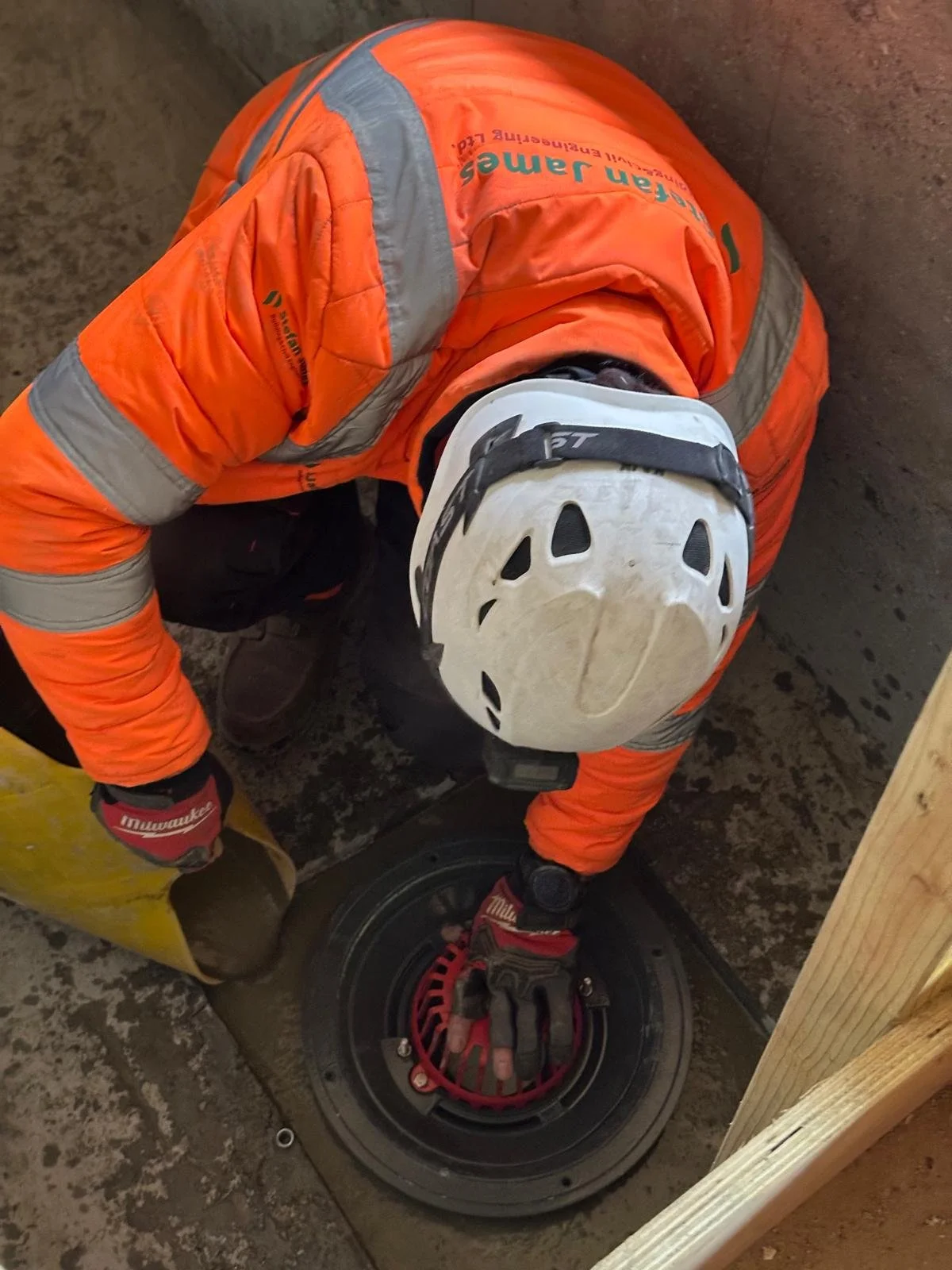 A worker in safety gear, including an orange jacket and a white helmet, is crouched down working on a circular utility access cover on the ground.