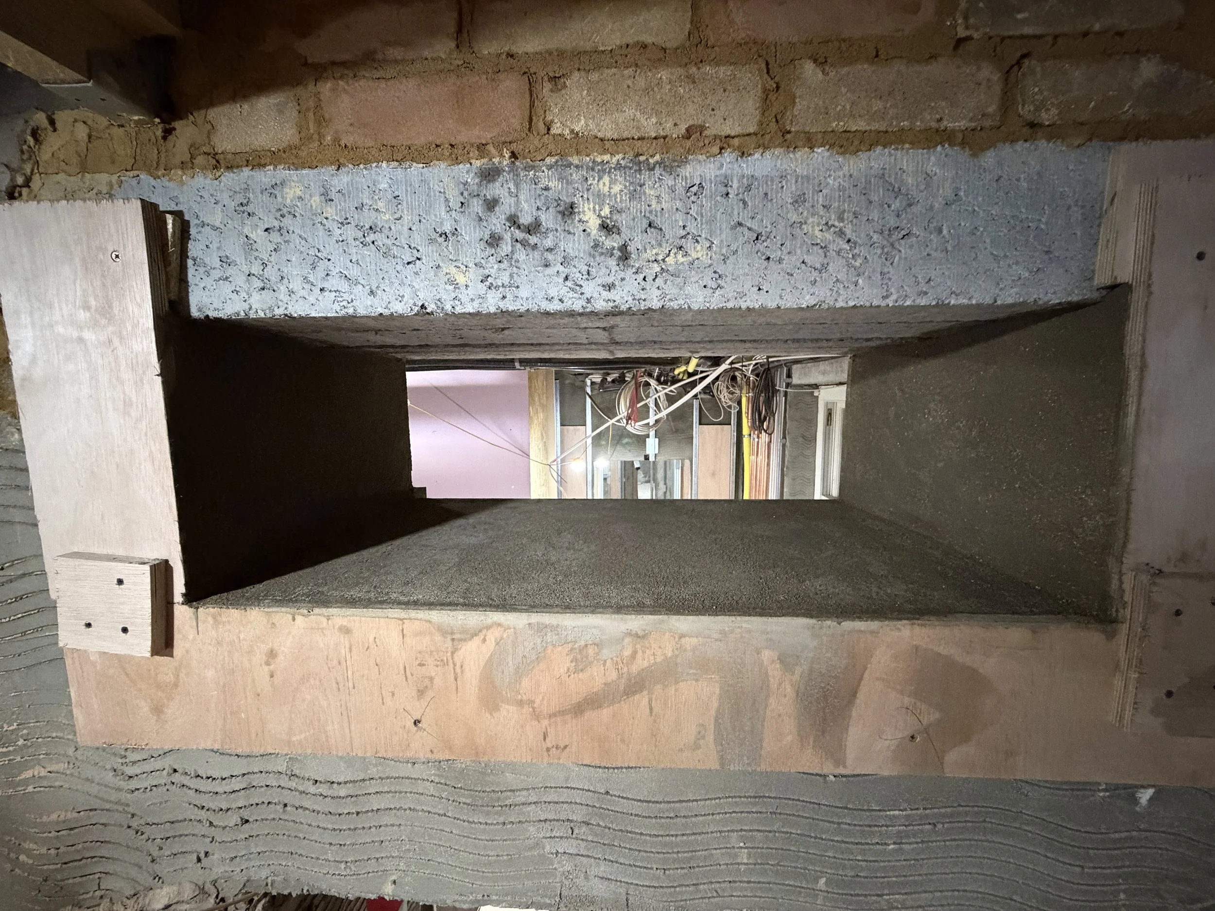 An unfinished construction site showing a rectangular concrete opening in a stud wall, with electrical wiring and insulation visible behind it, and brickwork above.