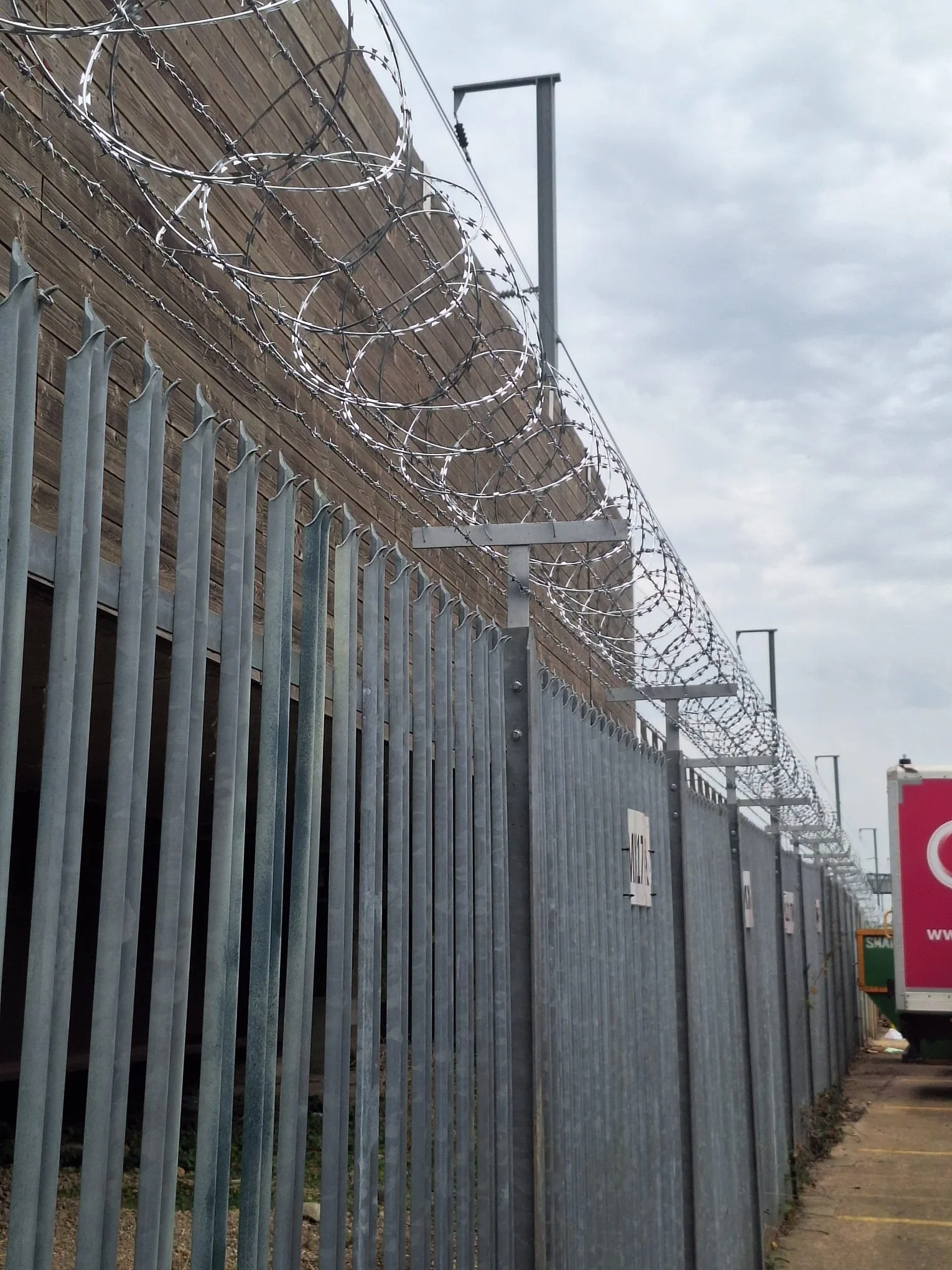 A tall metal security fence with pointed tips, topped with coiled barbed wire, running alongside a wooden wall with additional barbed wire, under an overcast sky. Streetlights and signs are visible in the background.