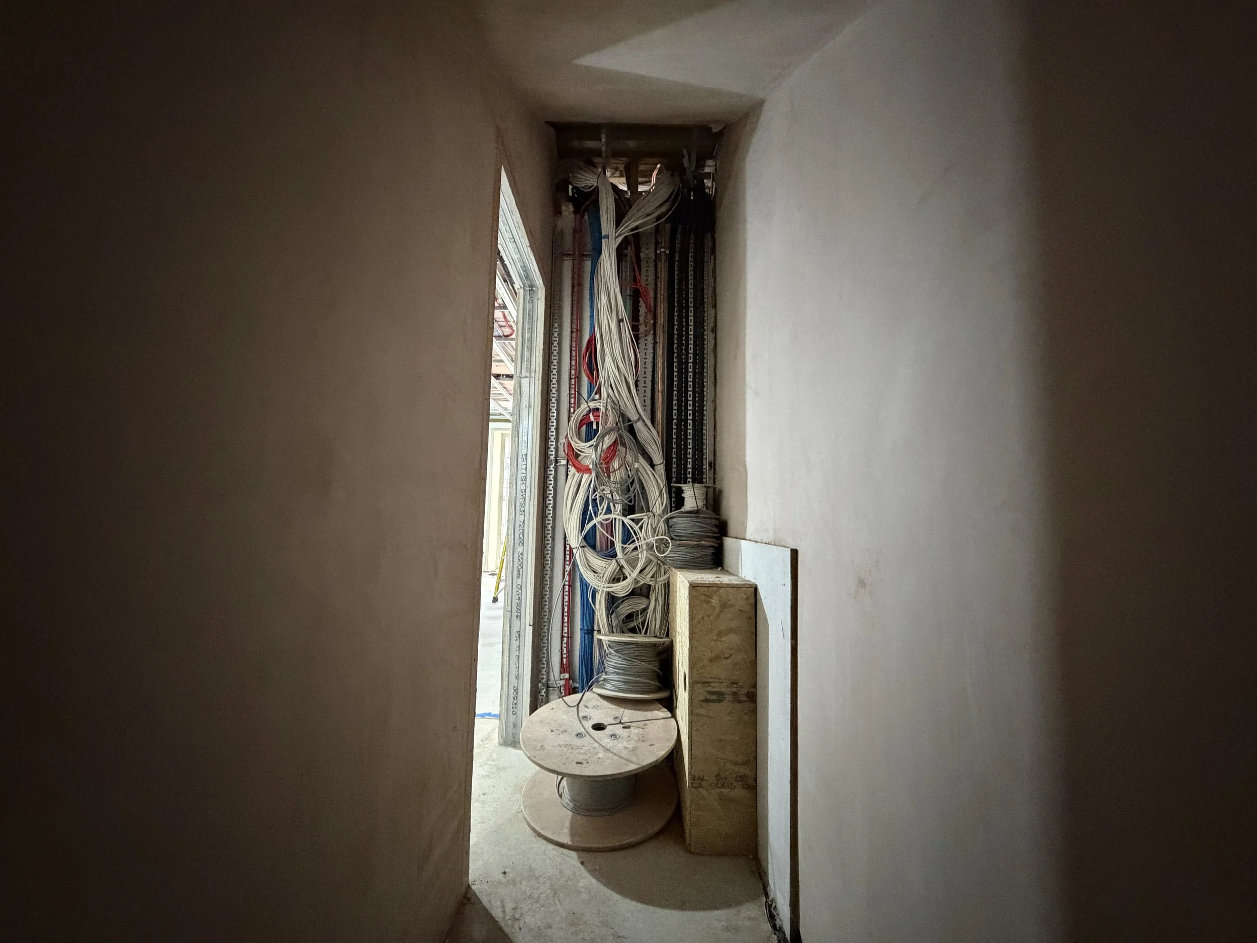 The image shows an unfinished room under construction with electrical wiring and conduit pipes installed. There is a window on the left side, and some construction materials are visible, including a reels of wiring and electrical panels.