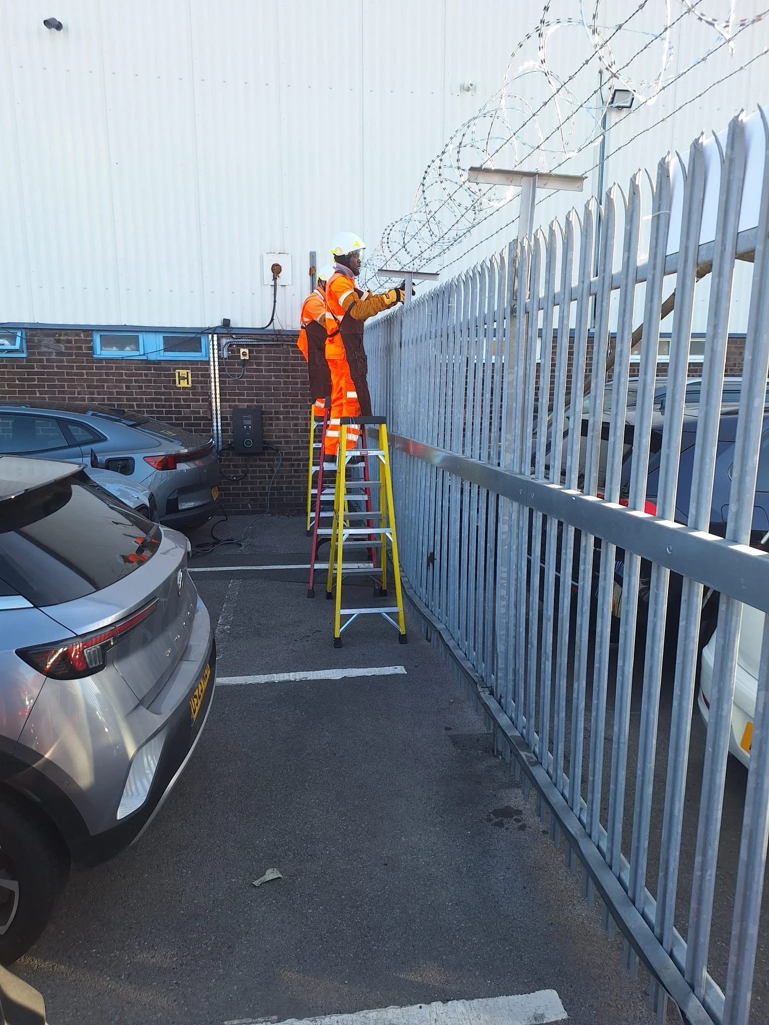Two workers in orange safety gear and white helmets installing barbed wire on top of a tall metal security fence outside a building with parking spaces and cars.