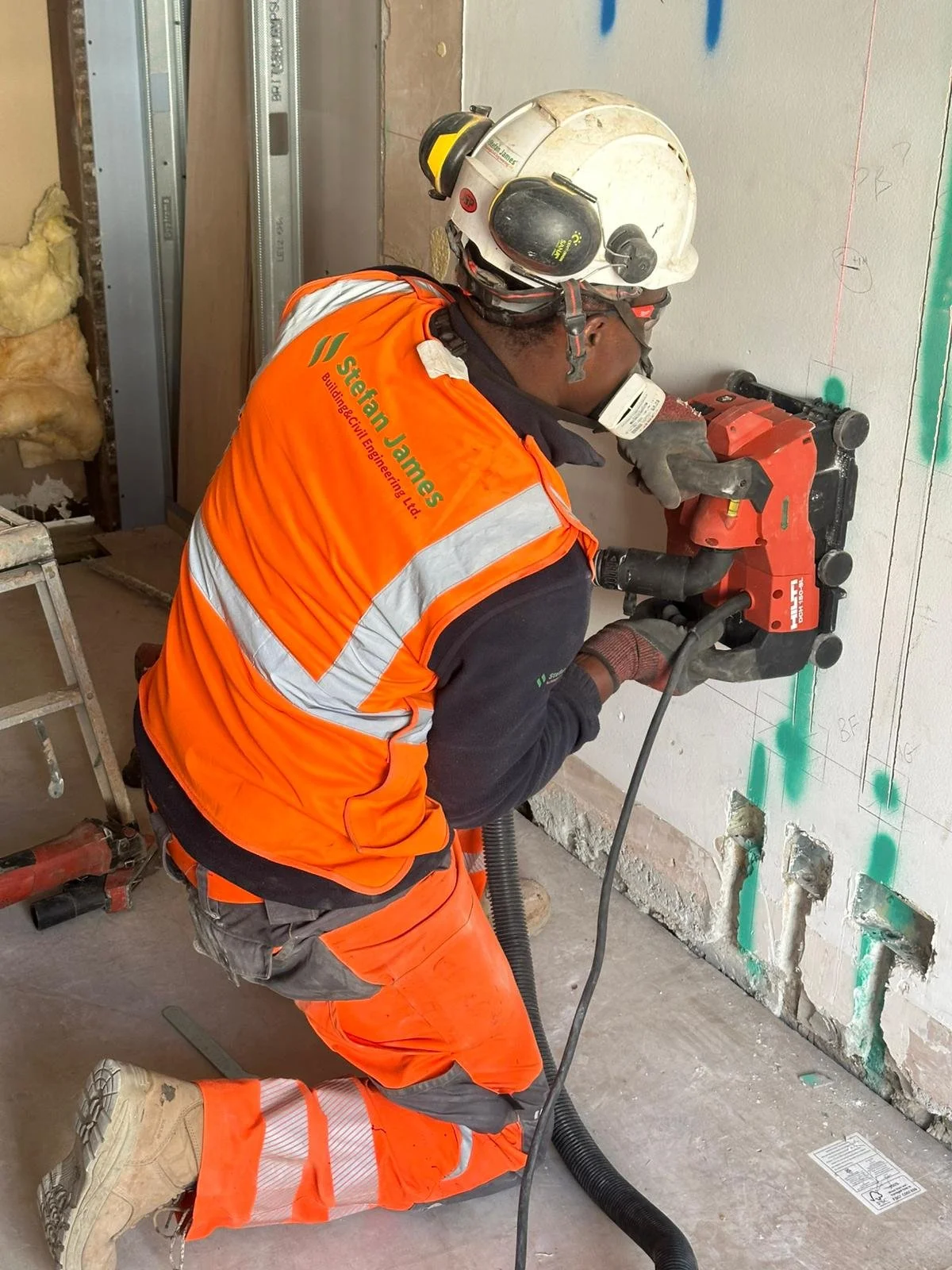 A construction worker using a jackhammer to break through a concrete wall. The worker is wearing an orange safety vest and a white helmet with ear protection.