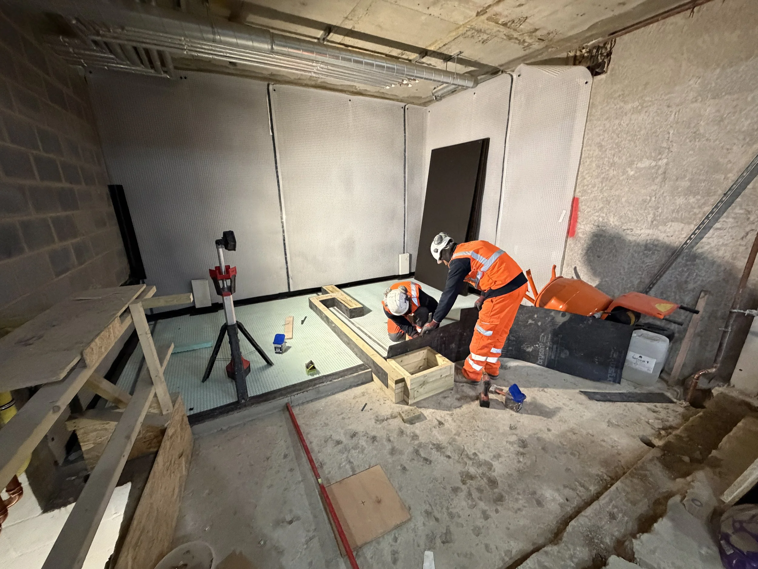 Workers in orange safety vests and helmets are installing flooring panels on a concrete surface in an unfinished basement or industrial space. Construction materials and tools are scattered around.