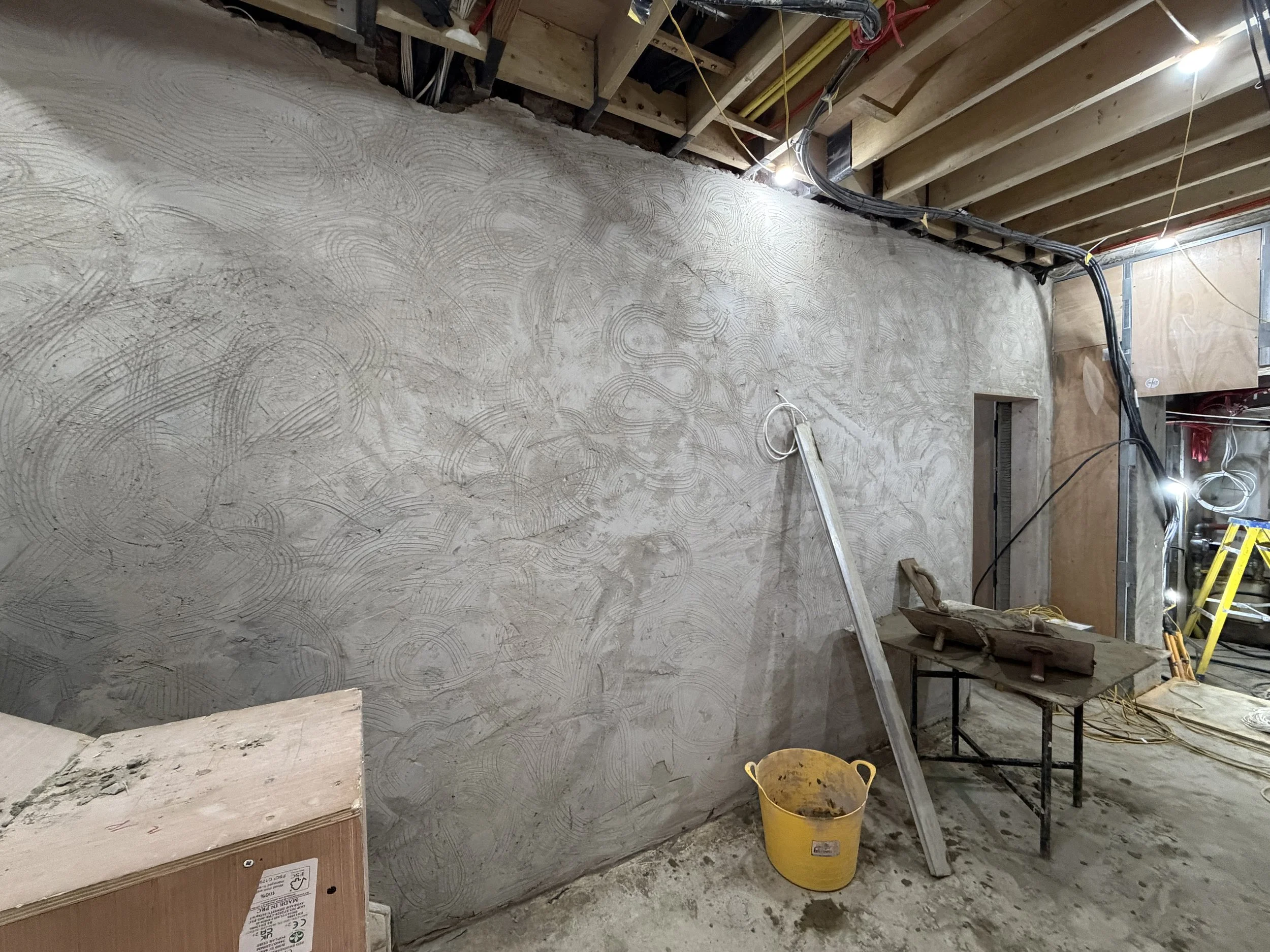 Interior construction site showing an unfinished room with electrical wiring, a partially installed wall, construction materials, a step ladder, and a yellow bucket.
