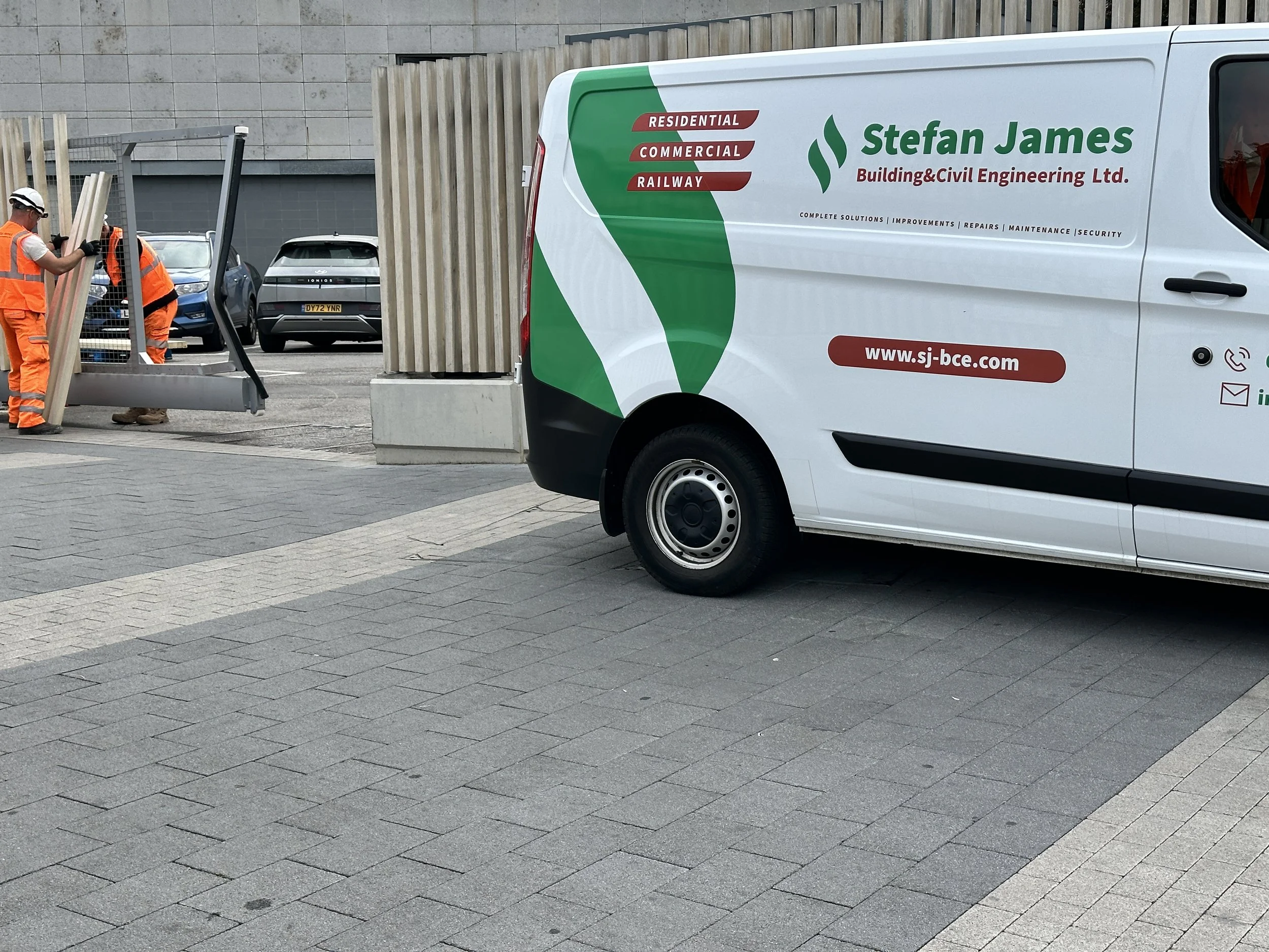 Construction workers in safety vests and helmets working near a white service van with company branding, on a paved outdoor area surrounded by a concrete wall.