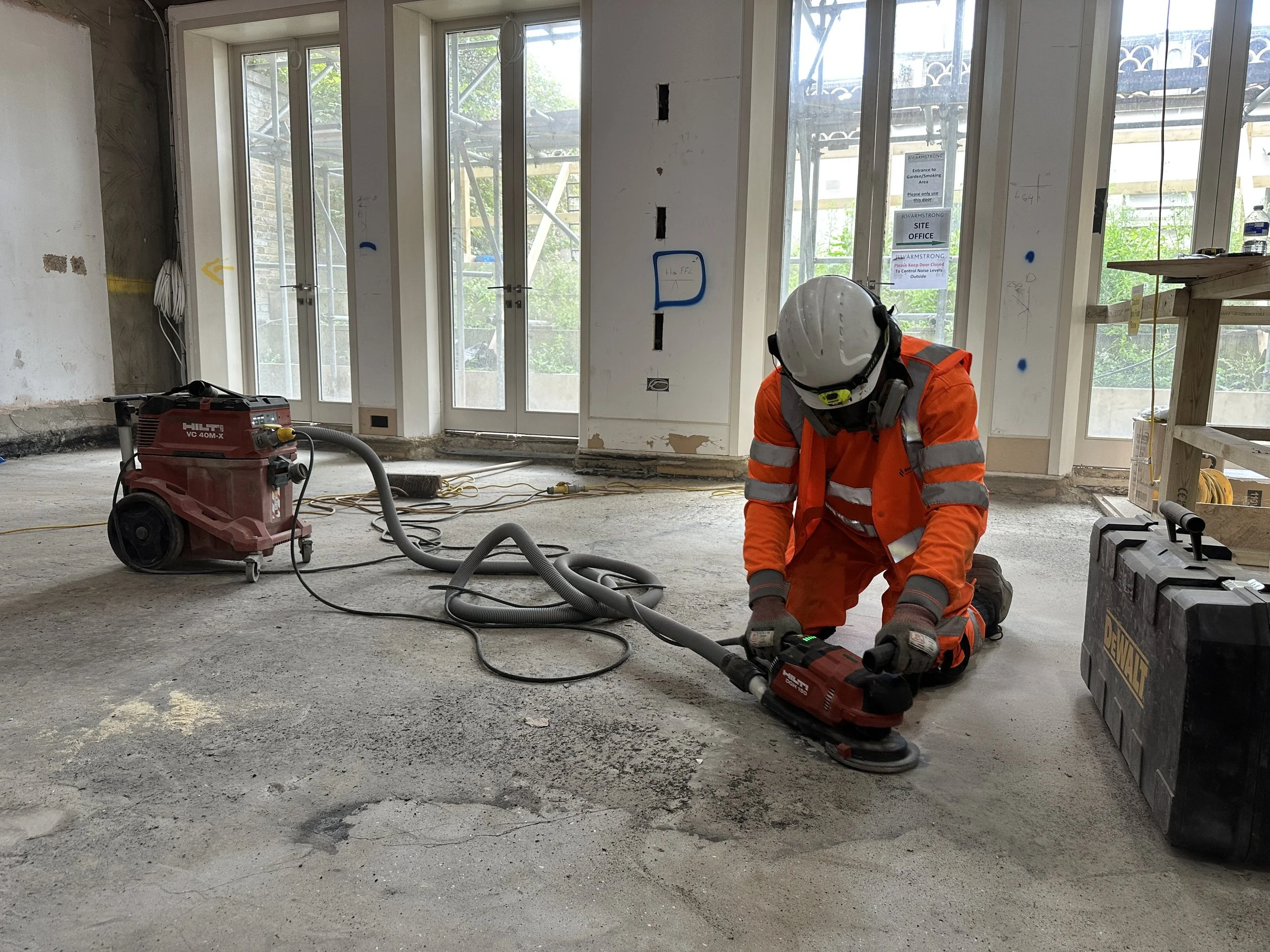 Construction worker in orange safety gear and white hard hat kneeling on concrete floor inside building under construction, working with a red Hilti power tool, with large windows showing scaffolding outside.