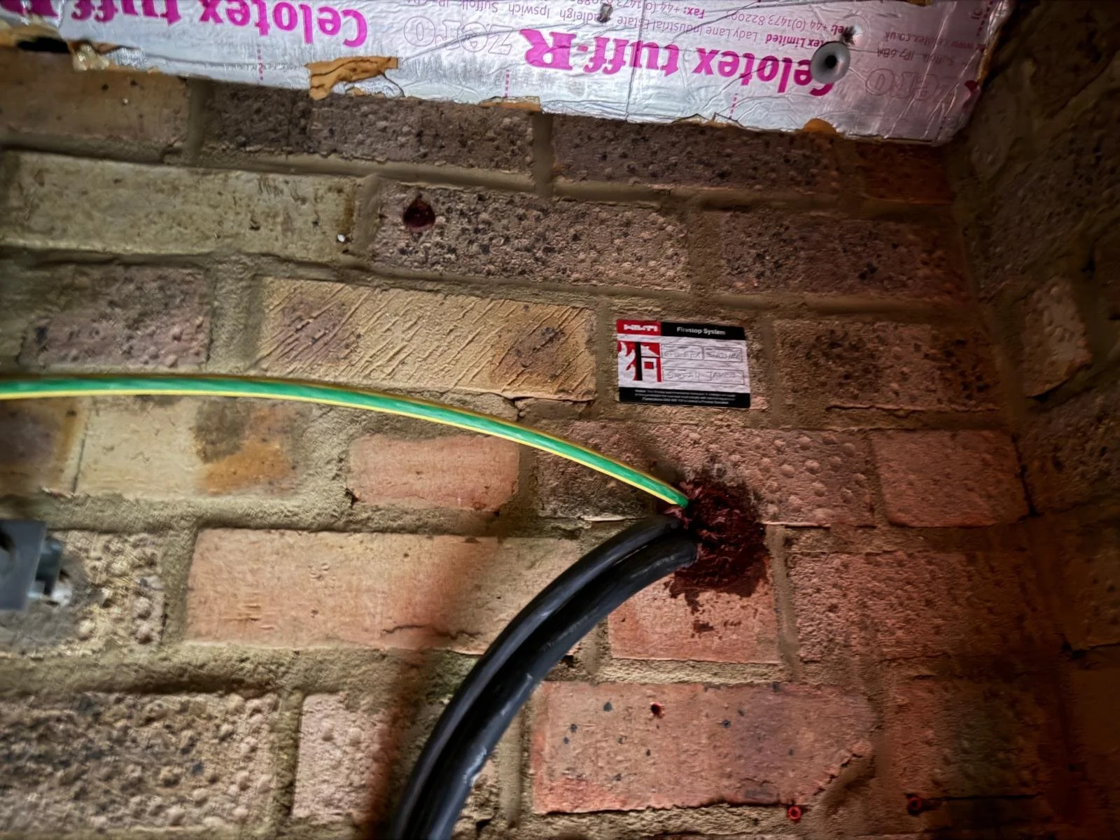 Close-up of an interior brick wall with a black electrical cable, a yellow-green wire, and two warning labels, indicating electrical work or wiring.