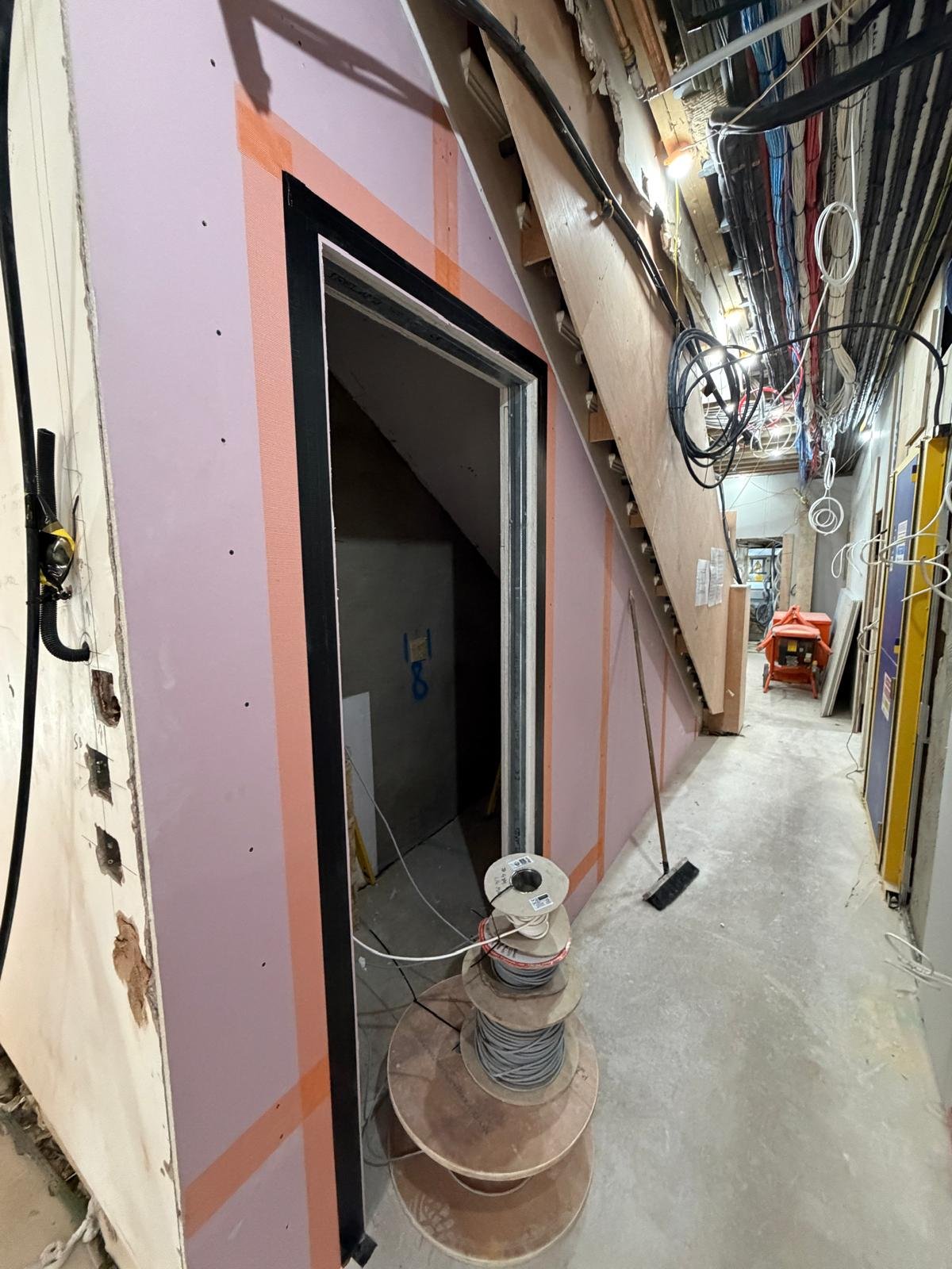 Construction site showing unfinished wall framing with insulation, wiring, and various construction tools including a broom, electrical cords, and a small red table in the background.