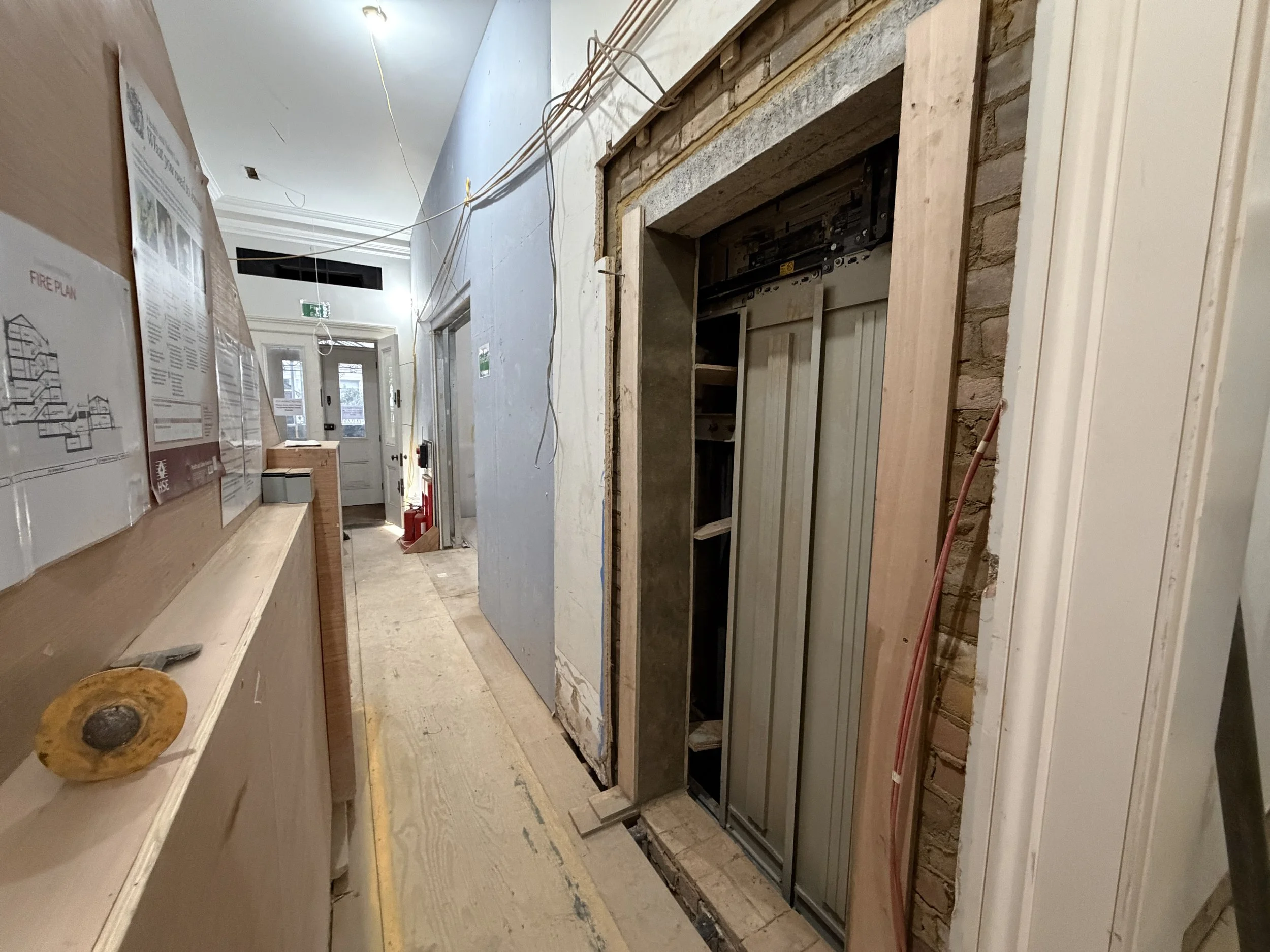 Interior of a building under construction, showing exposed brick and wood framing around an elevator shaft, with wiring hanging from the ceiling and construction plans posted on a wall.
