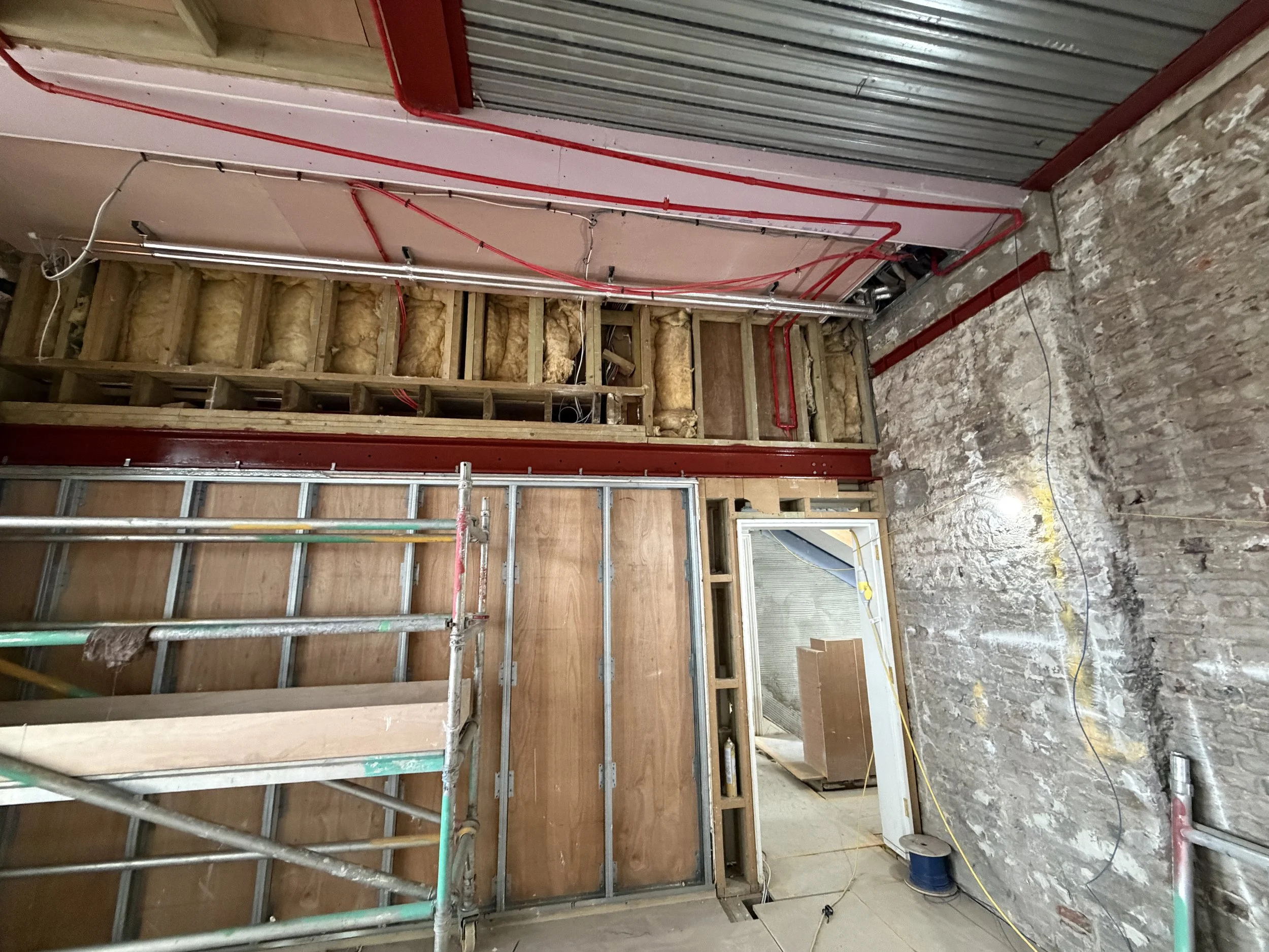Construction site showing a partially finished wall with insulation and metal framing, a window, and exposed brick on one side.