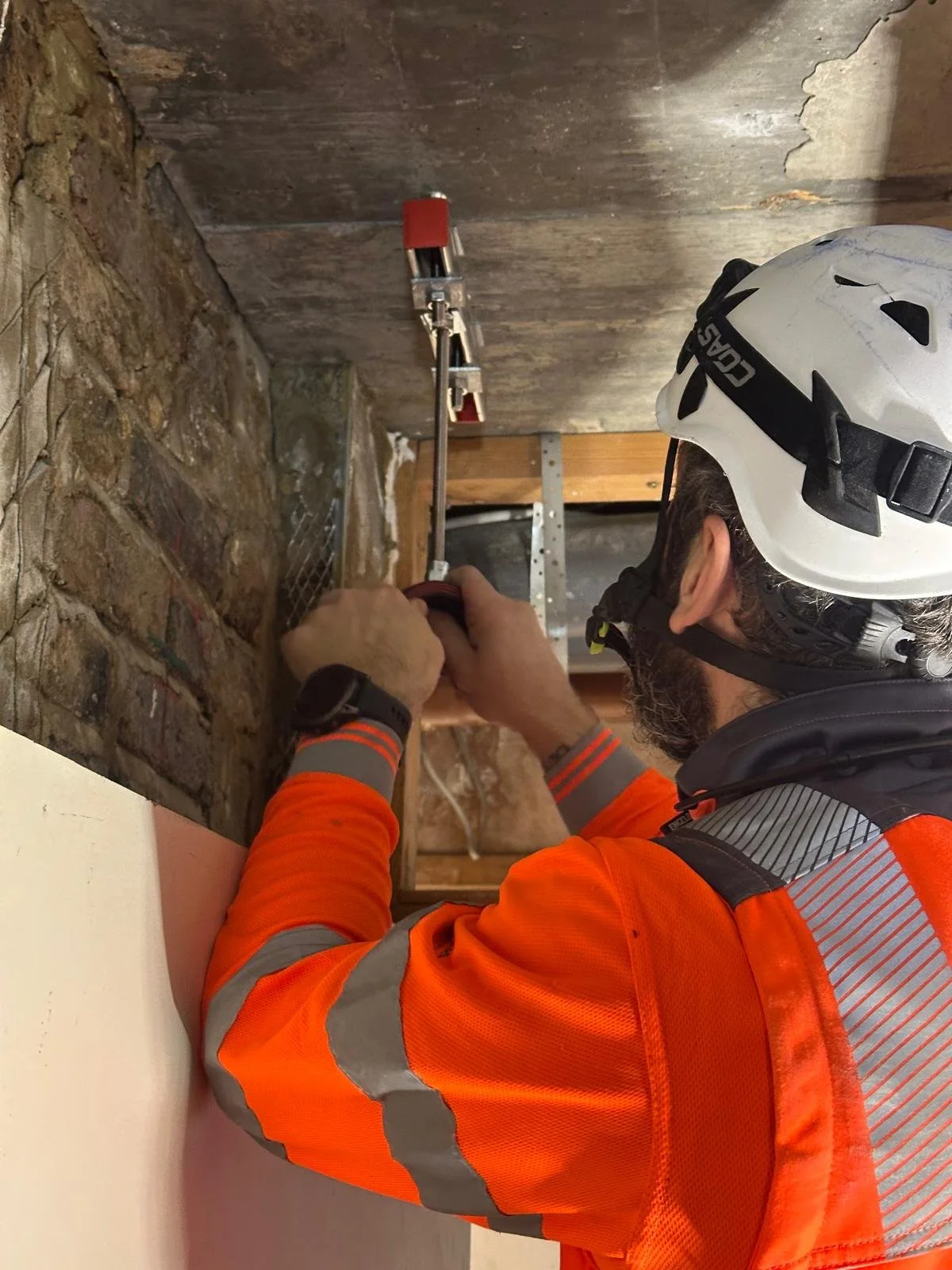A construction worker wearing an orange safety jacket and a white helmet is using a tool on a brick wall in what appears to be a building or renovation site.