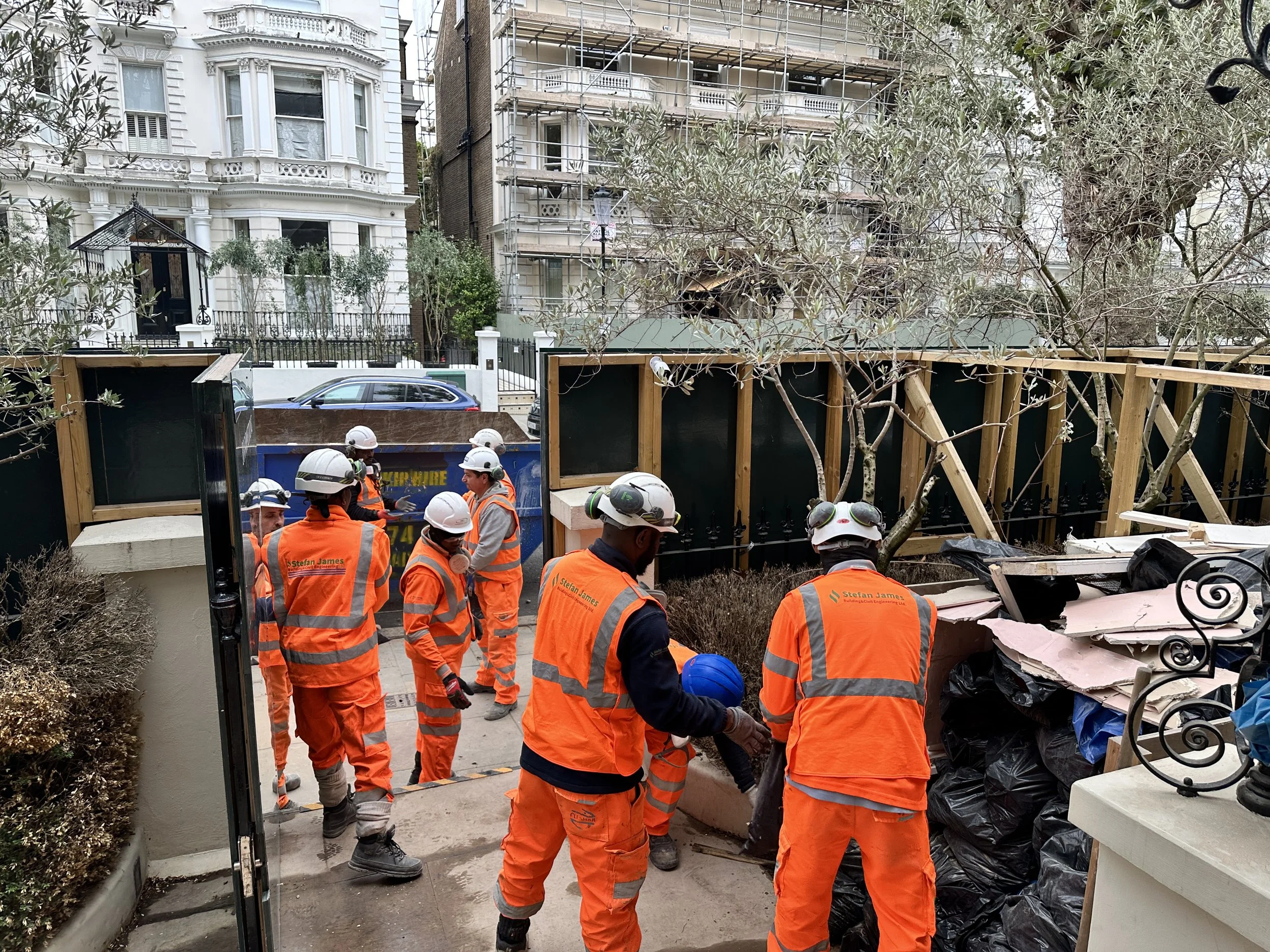 Construction workers wearing orange safety vests and white helmets are gathered near a construction site with wooden framing and debris. In the background, residential buildings are visible, one with scaffolding and another that is completed with ornate white architecture.