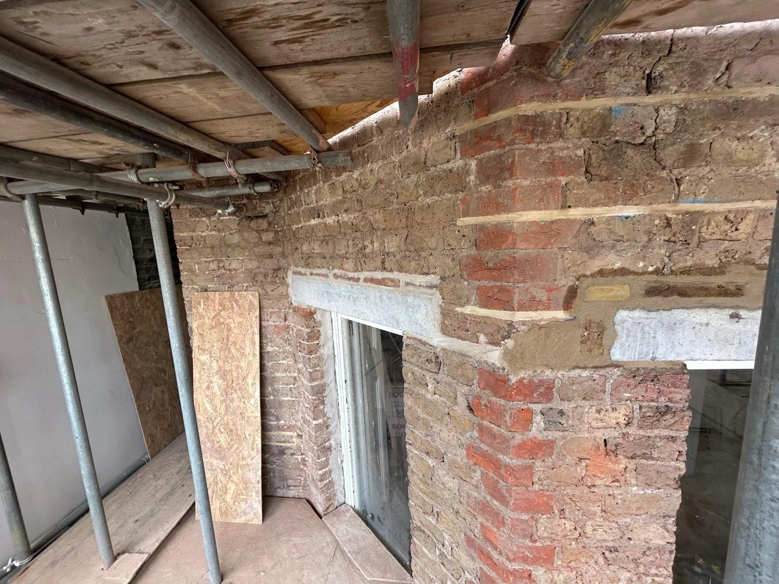 A close-up of a brick wall under renovation with exposed mortar, with scaffolding and wooden planks at the construction site.