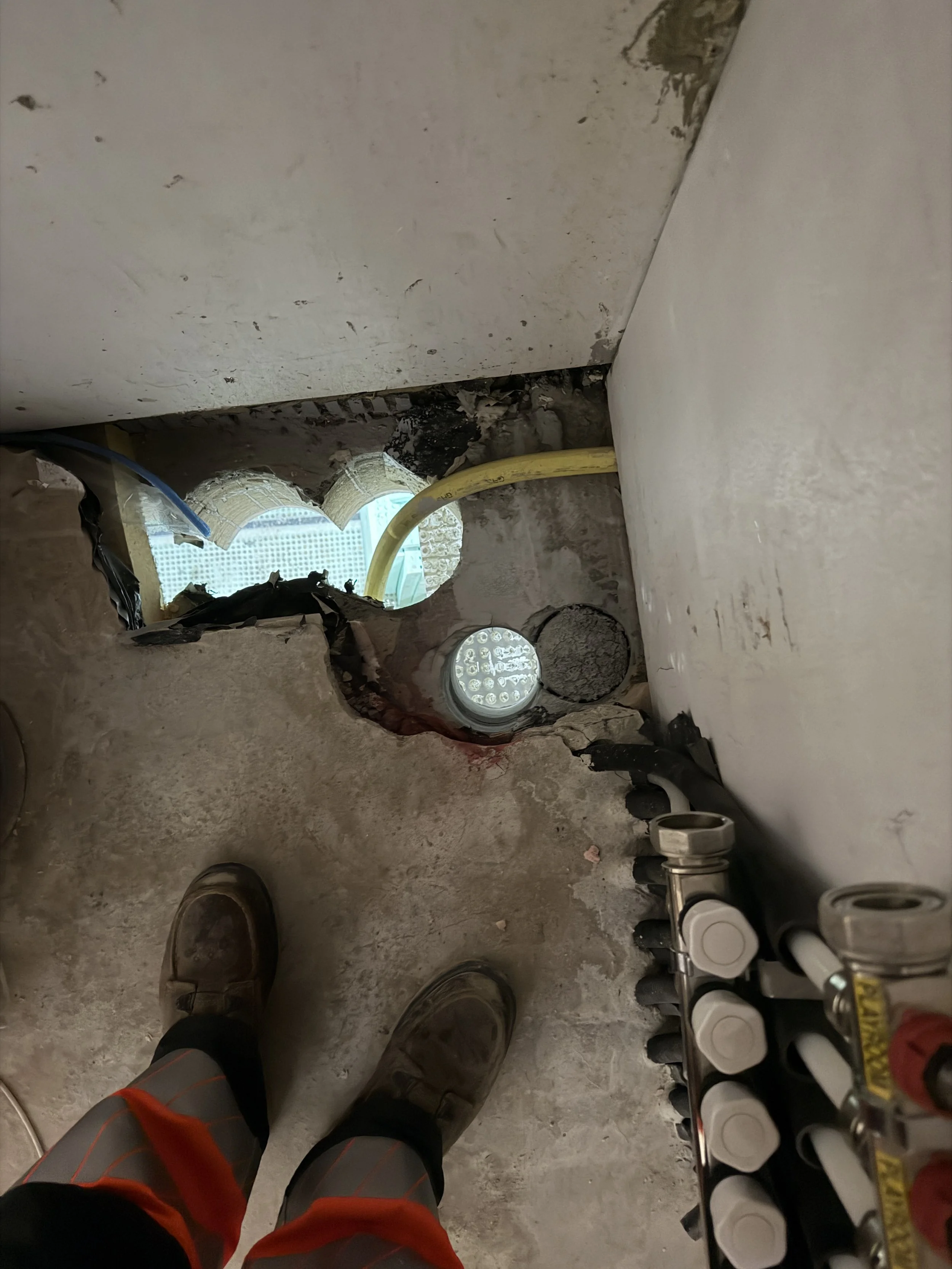 Person standing on a concrete floor wearing work boots and orange safety pants, with plumbing or gas piping visible nearby.