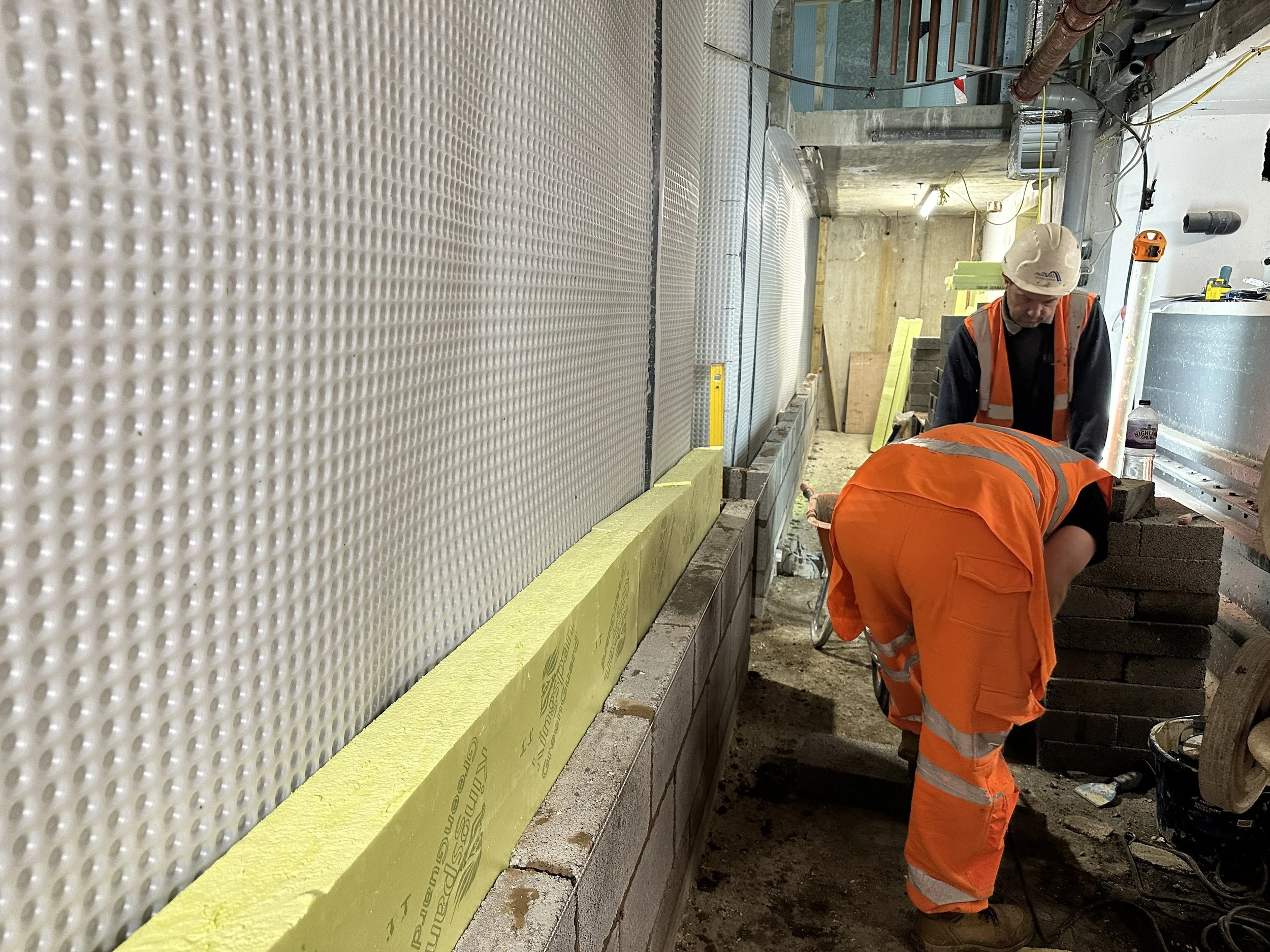 Construction workers wearing orange safety uniforms and helmets working on a building site. One worker is down, and another is standing, working near a partially built wall with insulation material and pipes.