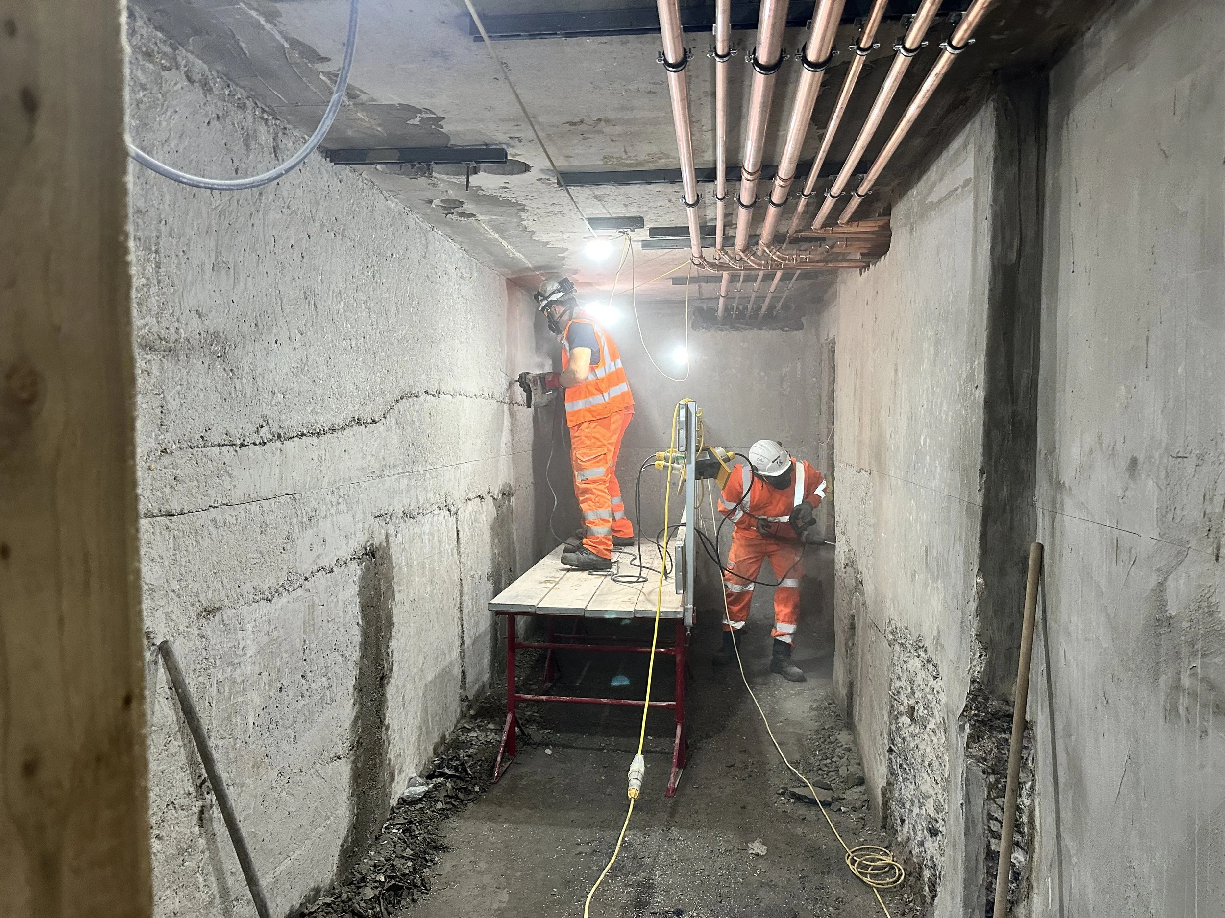Construction workers in orange safety uniforms and helmets working inside a concrete tunnel. Pipes are installed on the ceiling. Bright lights illuminate the workspace.