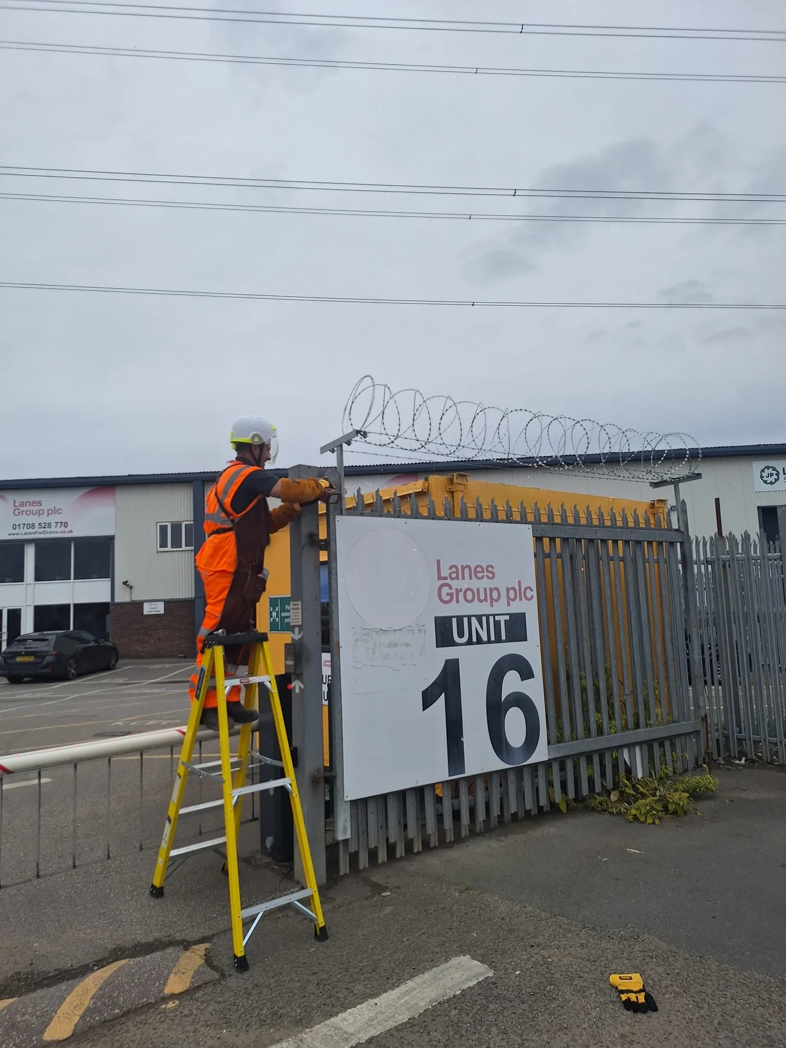 A worker in an orange safety vest and helmet standing on a yellow ladder, installing or maintaining a sign near a fenced area with contract barbed wire. The sign displays the name "Lanes Group plc" and the number 16. There are cars parked behind the fence and power lines above.