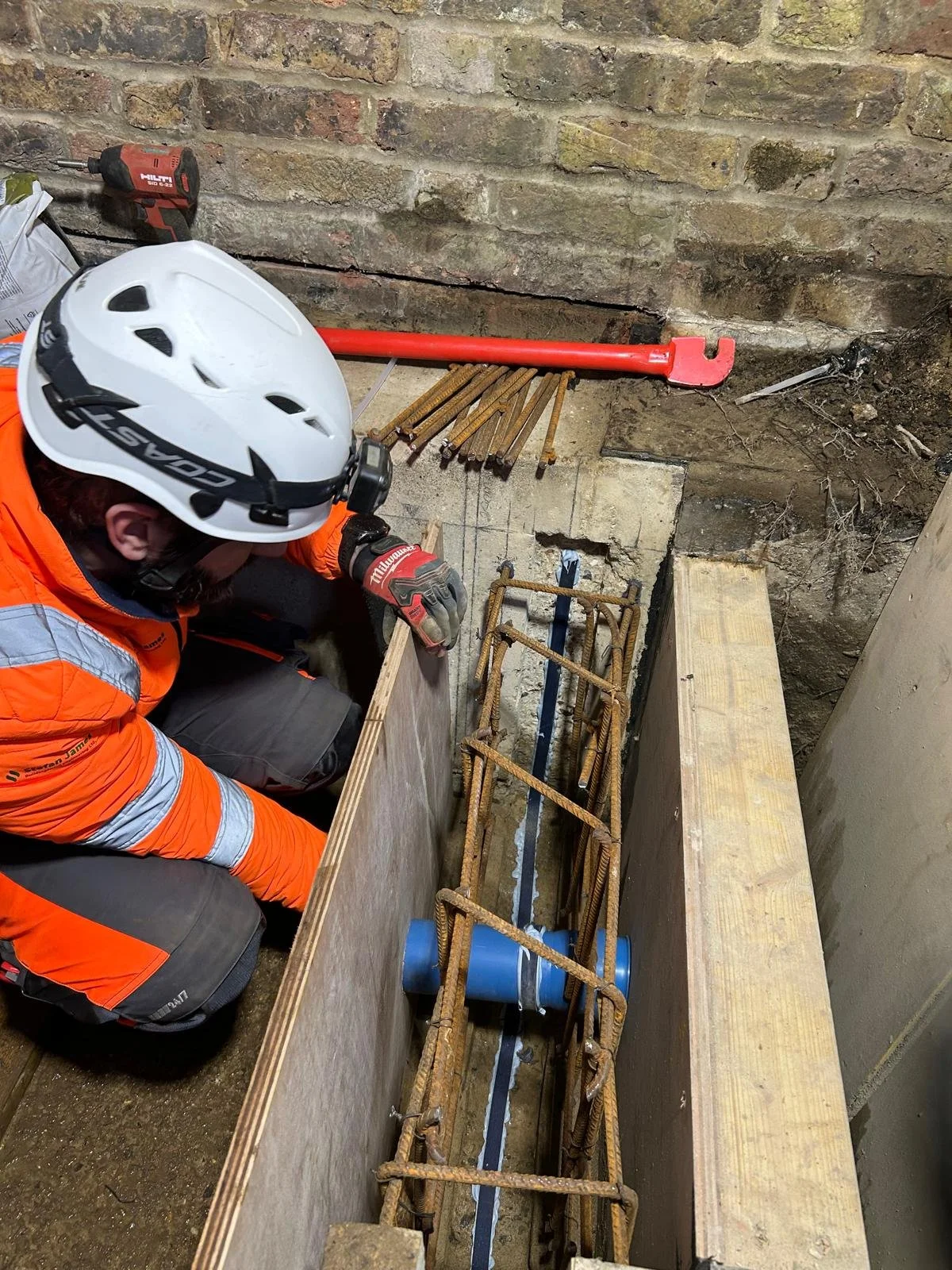 Construction worker in orange safety gear and a white helmet working on a renovation project in a basement or underground space, with exposed brick walls and construction tools nearby.