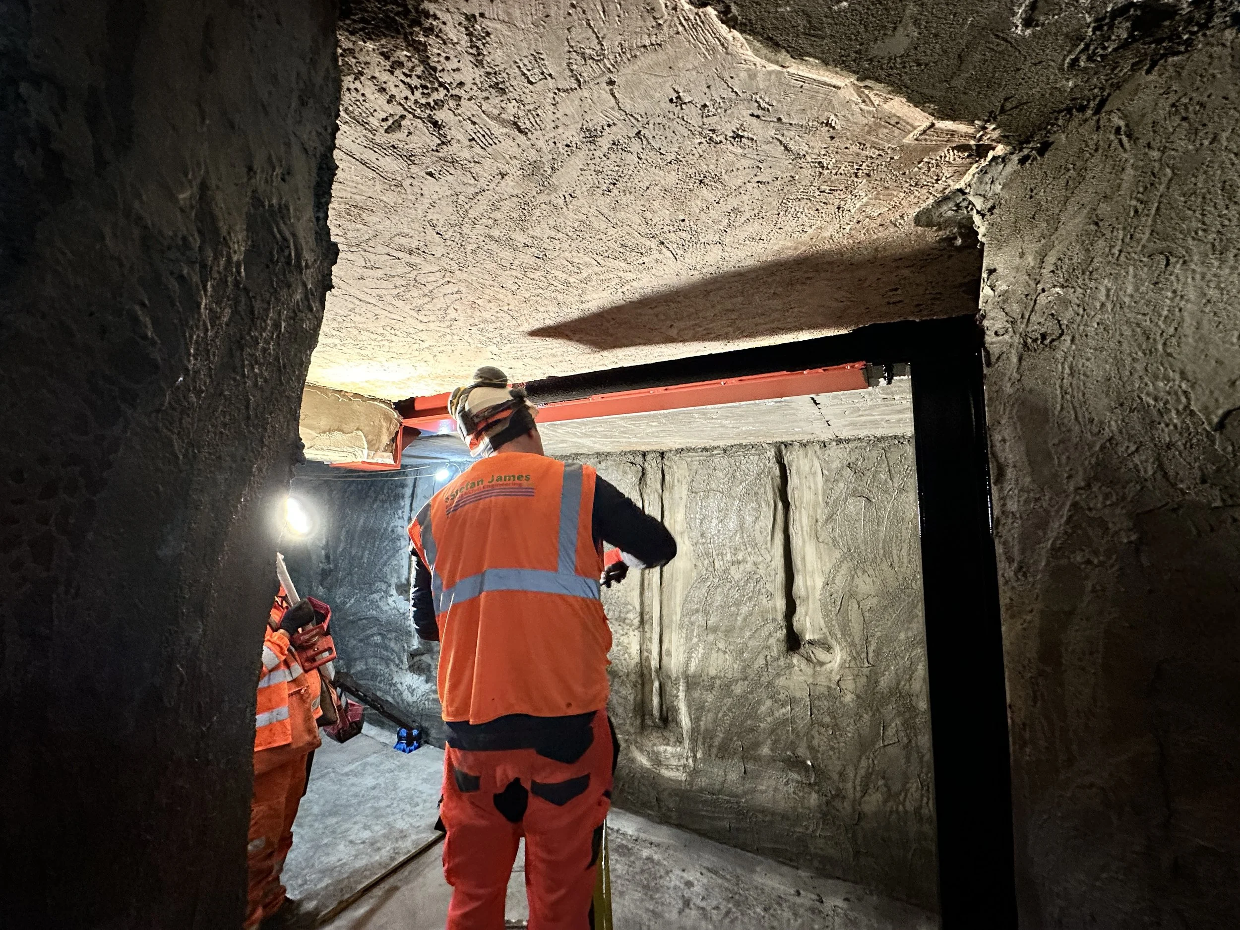 Construction workers in orange safety gear working on a large concrete wall inside a tunnel.