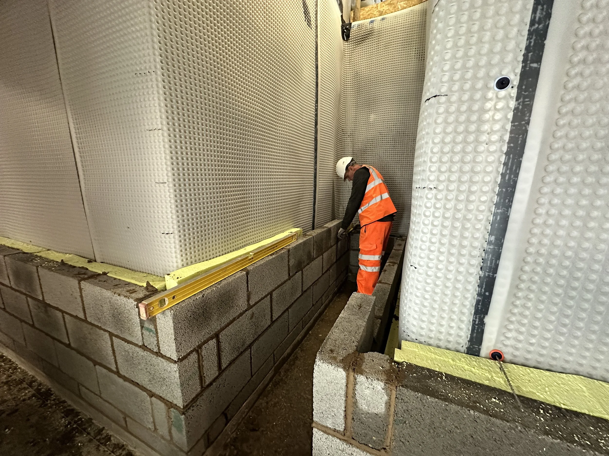 Construction worker in an orange safety vest and white hard hat working on a cement block wall, with white plastic sheeting on the right side of the image and a perforated metal wall behind him.