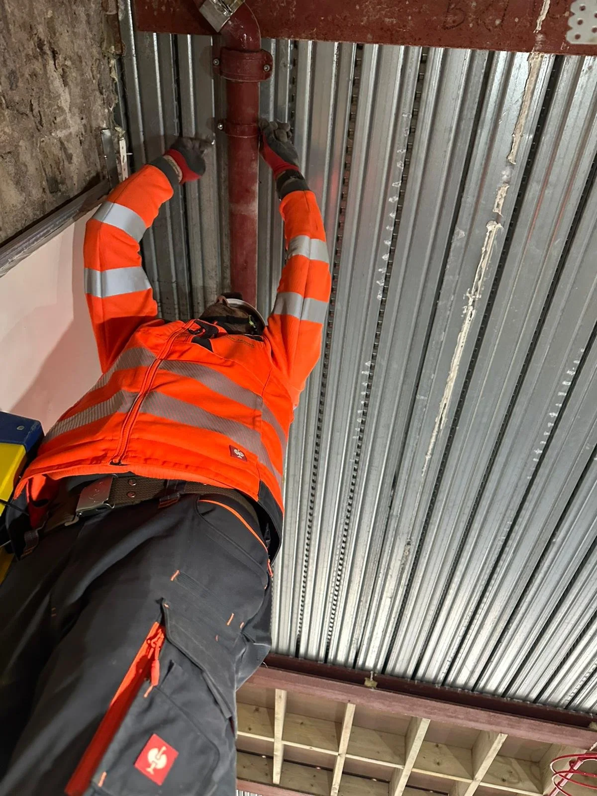 Worker in orange safety jacket and black pants installing or working on a red pipe attached to a metal ceiling inside a building under construction.
