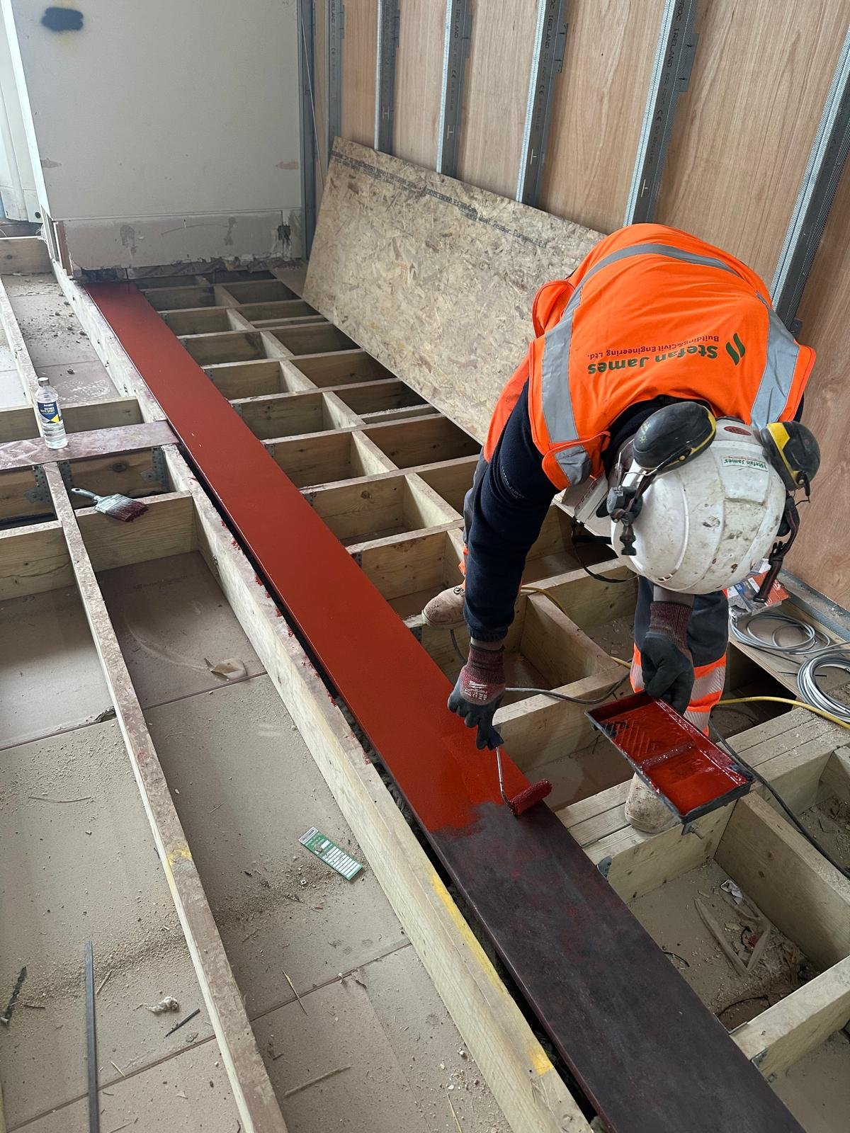 A construction worker painting a wooden floor frame with red paint inside a building under construction. The worker is wearing a white safety helmet, orange safety vest, dark gloves, and long black pants. Tools and materials are scattered around.