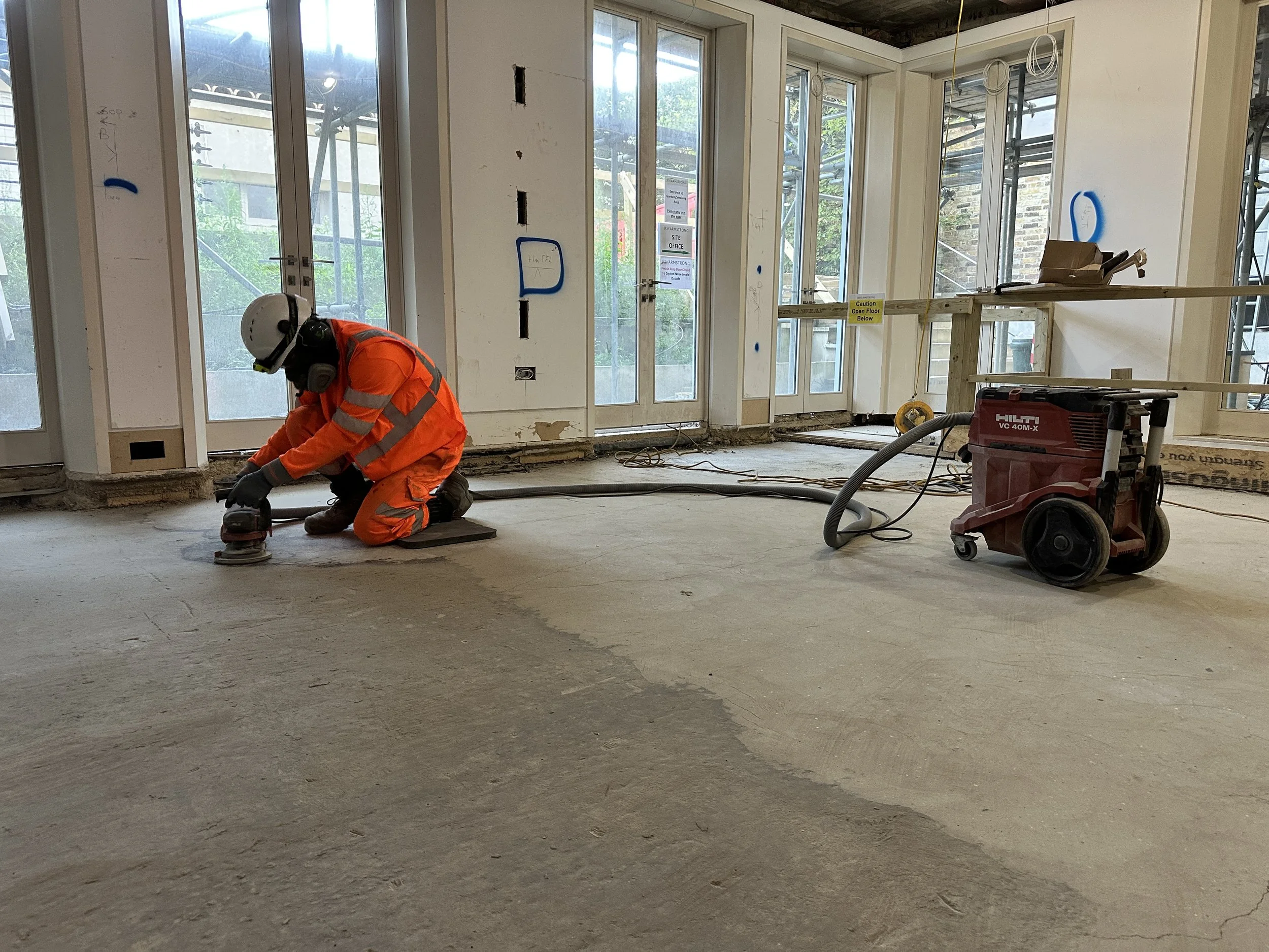 Construction worker wearing orange safety gear, kneeling on floor, working on construction site indoors with large glass doors and windows, construction tools and equipment around.