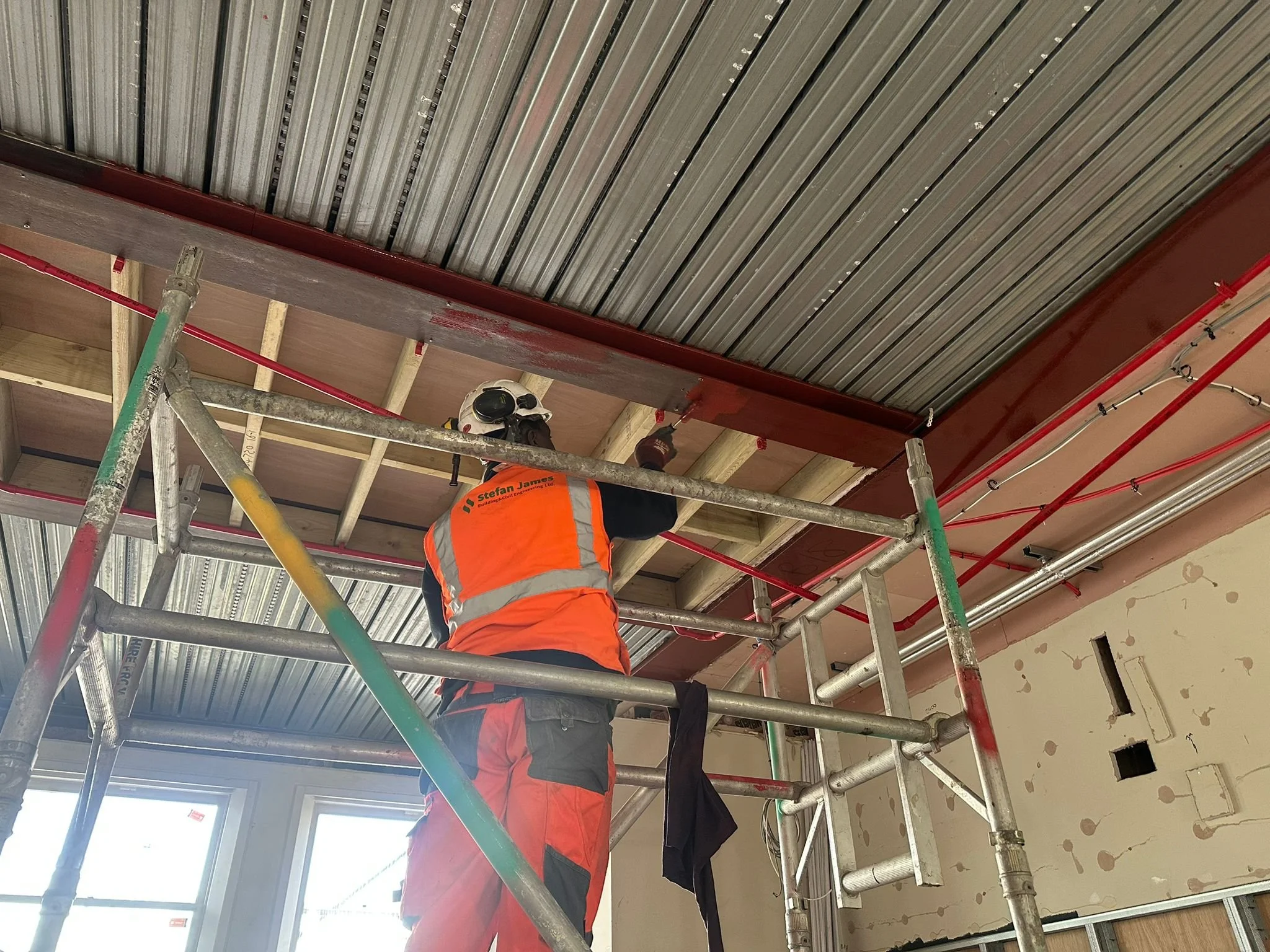 A construction worker wearing an orange safety vest and a white hard hat standing on scaffolding inside a building under construction. The ceiling has metal beams and pipework.