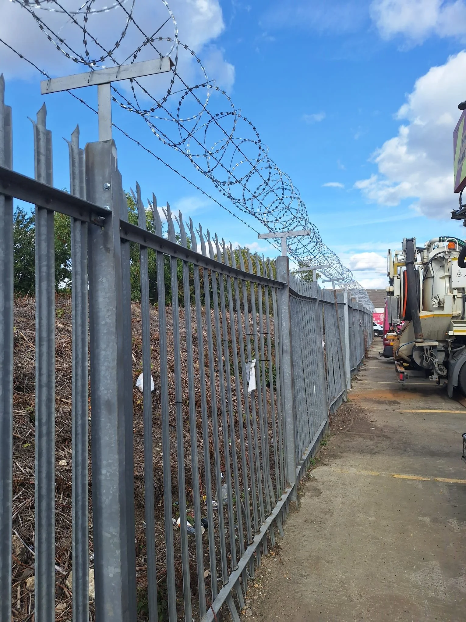 A tall metal fence topped with barbed wire coils runs along a sidewalk. Behind the fence, there is a slope covered with dry leaves and vegetation. To the right, a utility vehicle is parked on the paved area. The sky is partly cloudy with blue sky visible.