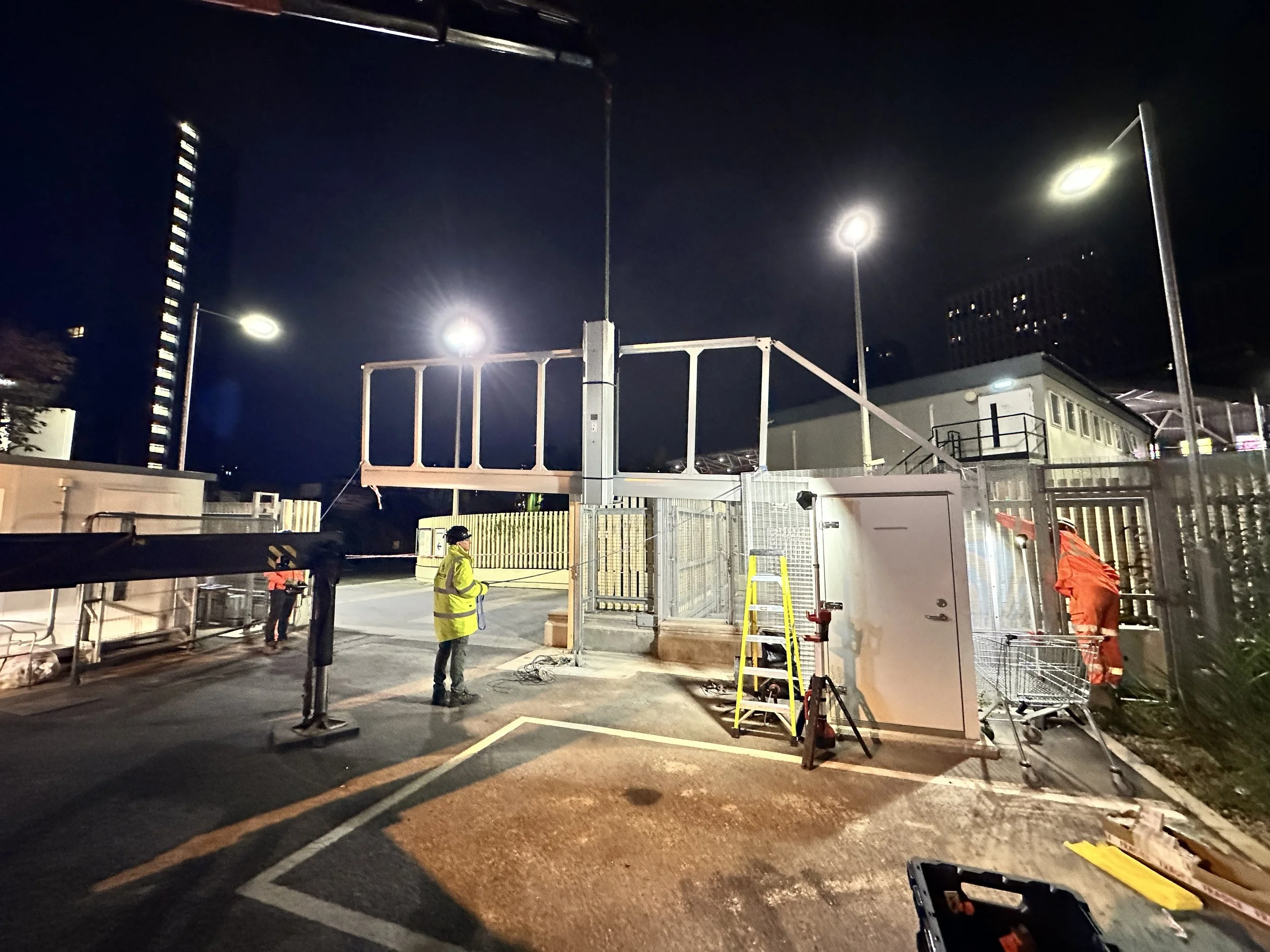 Nighttime scene of a construction or maintenance site with workers, equipment, and overhead lighting in an outdoor parking lot or similar area.