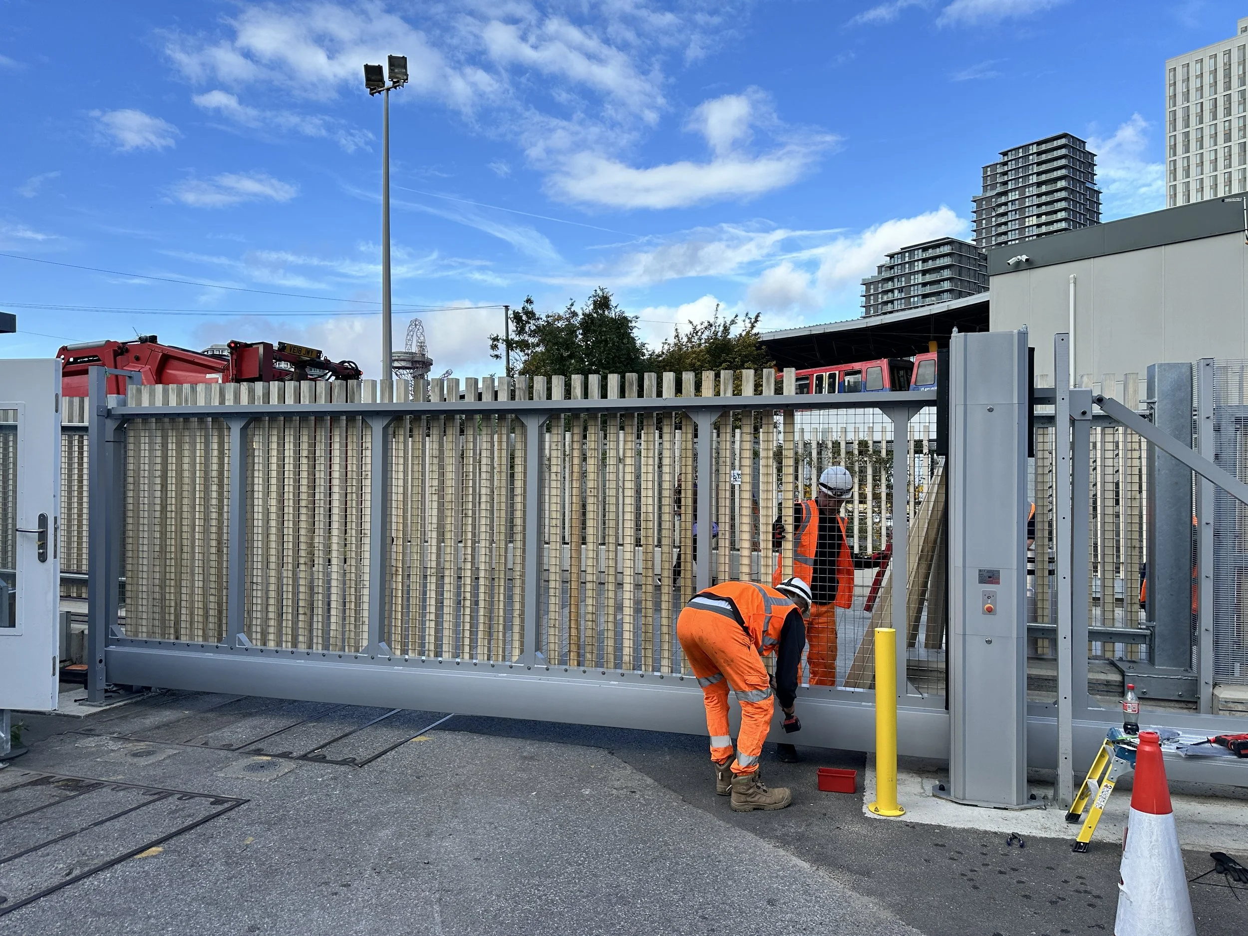 Construction workers in orange safety uniforms installing a sliding gate at a train station or similar transportation facility, with skyscrapers in the background and a blue sky with clouds overhead.