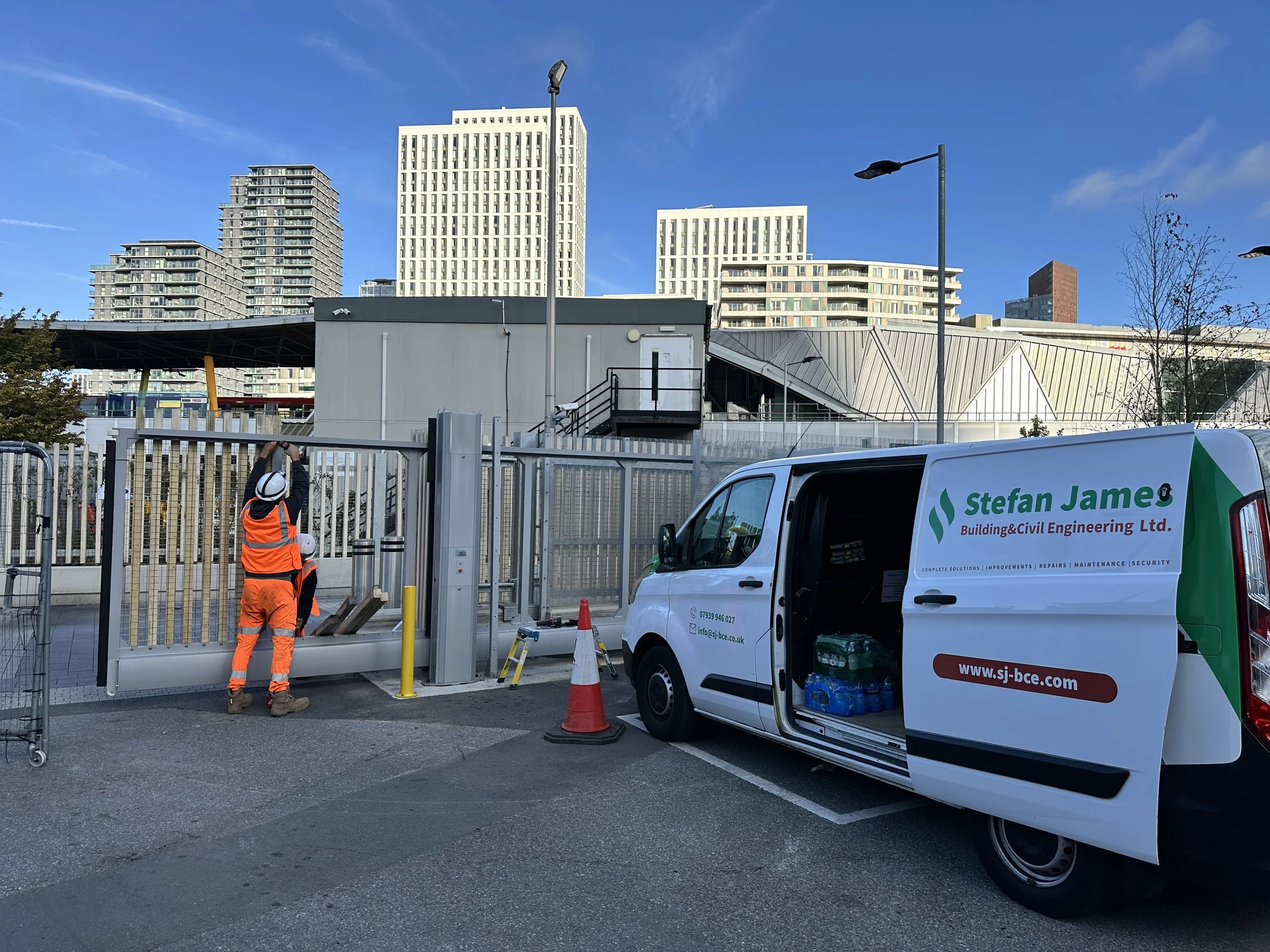 Construction workers in orange safety vests and helmets working near a white vehicle with company branding, on a city street with high-rise buildings in the background.