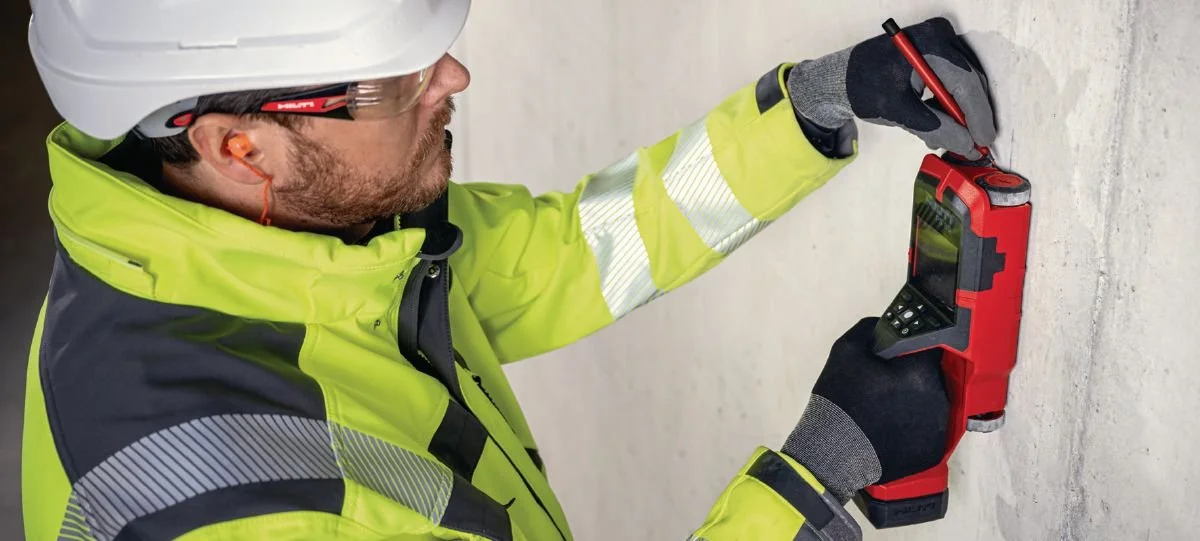 Construction worker in a hard hat and high-visibility jacket uses a measuring device on a wall.