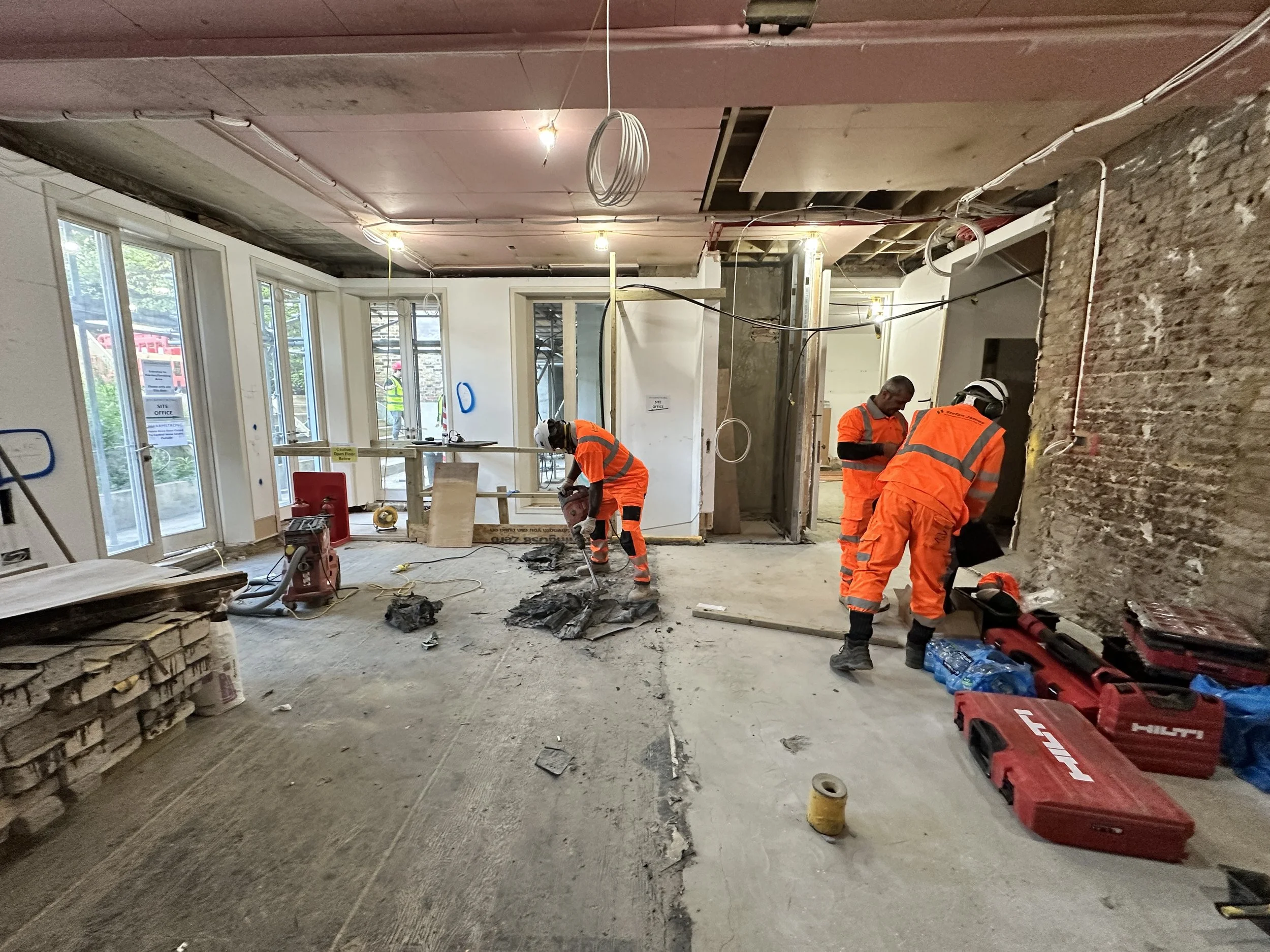 Construction workers in orange safety uniforms working inside a building under renovation with exposed walls, electrical wiring, and construction tools and materials around.