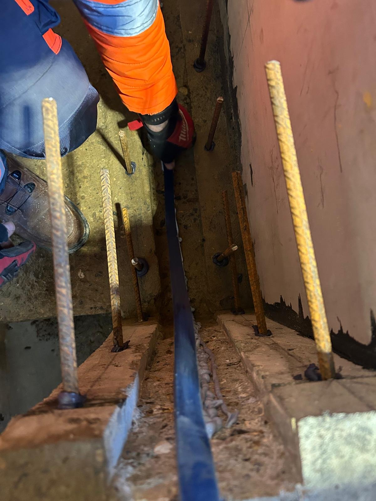 A construction worker wearing an orange safety jacket and black boots is working on a construction site. The photo is taken from above, showing the worker standing inside a narrow space reinforced with metal bars and wooden supports, with a concrete or drywall wall on one side.