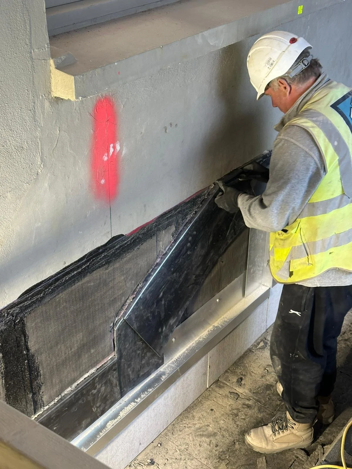 Construction worker wearing a white hard hat, yellow safety vest, and work boots, working on a construction project on a building wall with insulation and ductwork. A red spray-painted marker is on the wall.
