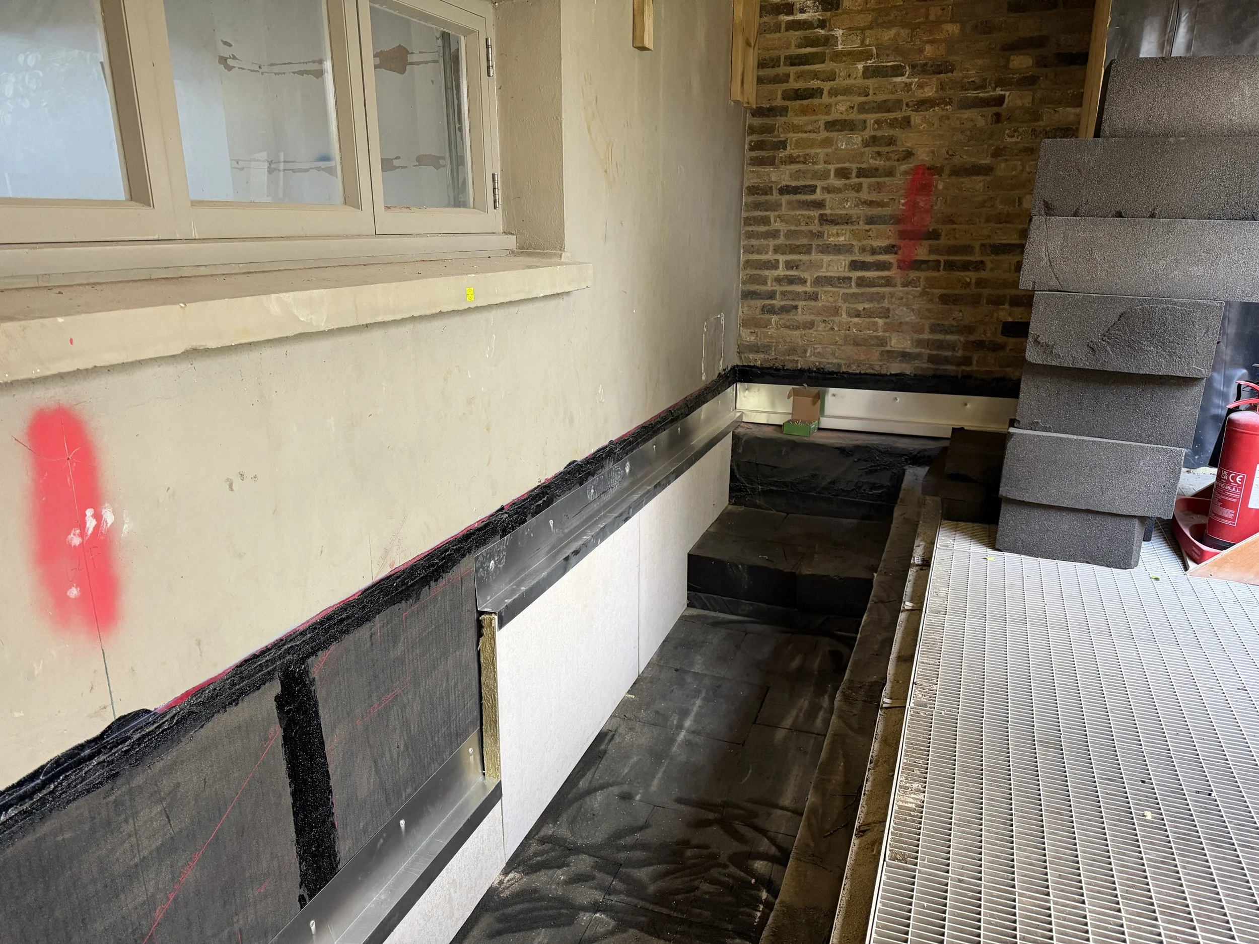 Interior view of a construction or renovation site with an unfinished wall featuring bricks and off-white paint, a window, and construction materials such as foam blocks and a fire extinguisher on the floor.