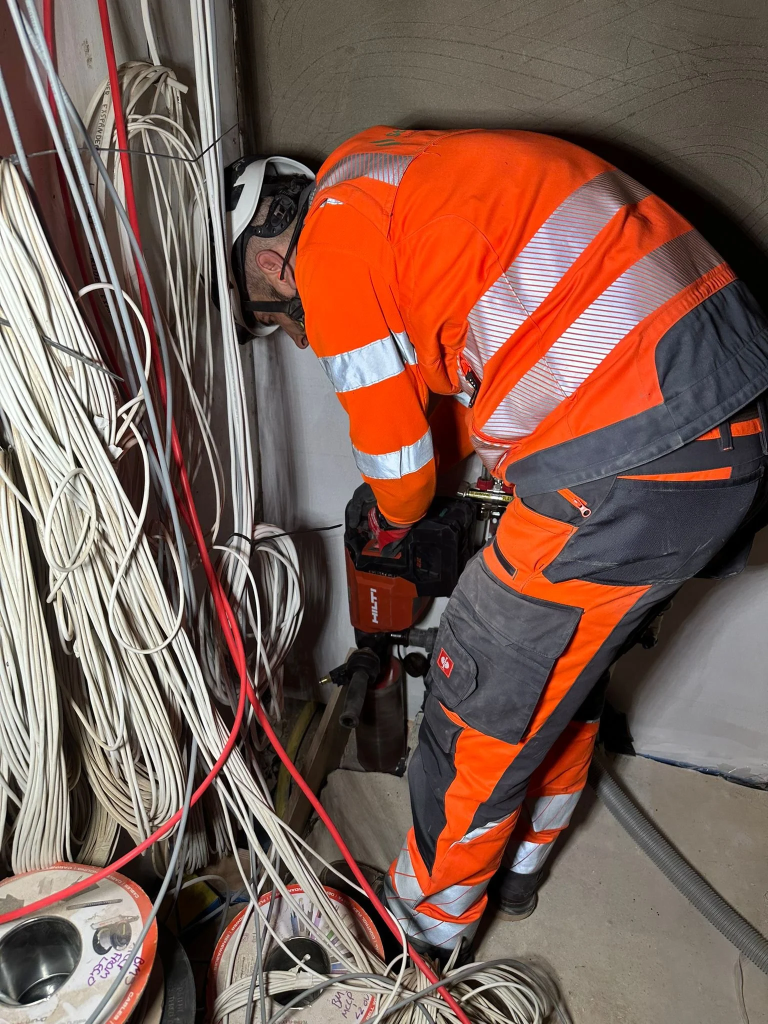 A worker in orange safety gear and a helmet working with tools among a tangled mass of white and red cables in an industrial or electrical setting.