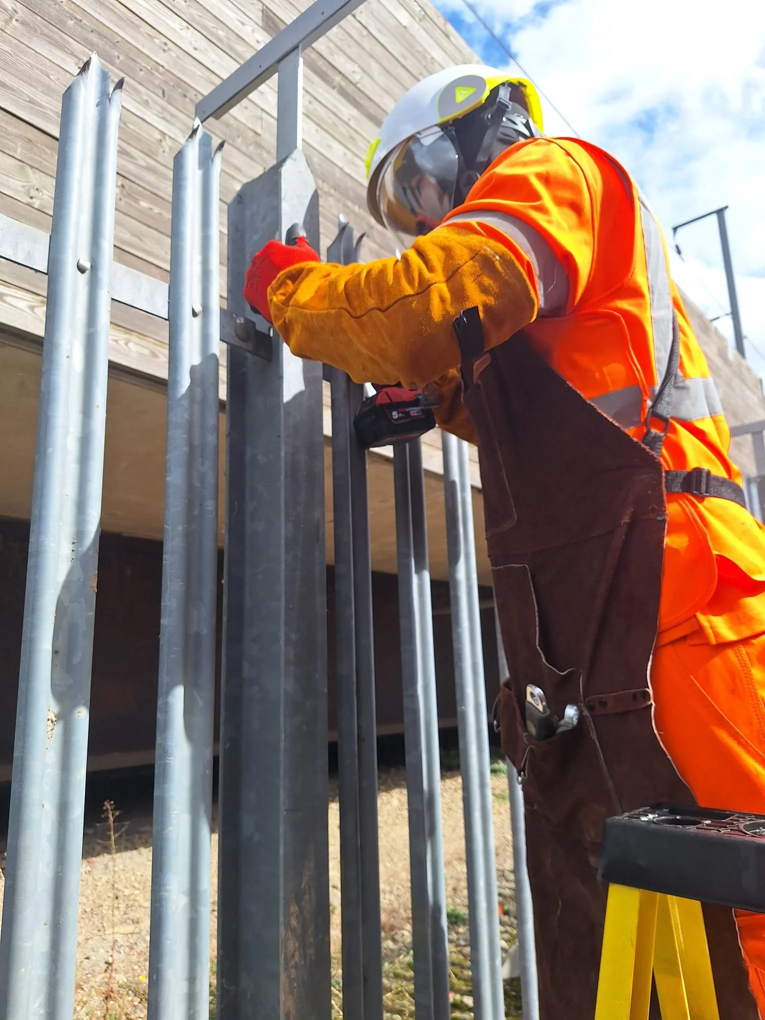 A construction worker wearing a white safety helmet, orange high-visibility vest, yellow gloves, and work gear, installing or securing metal fencing outside under a partly cloudy sky.