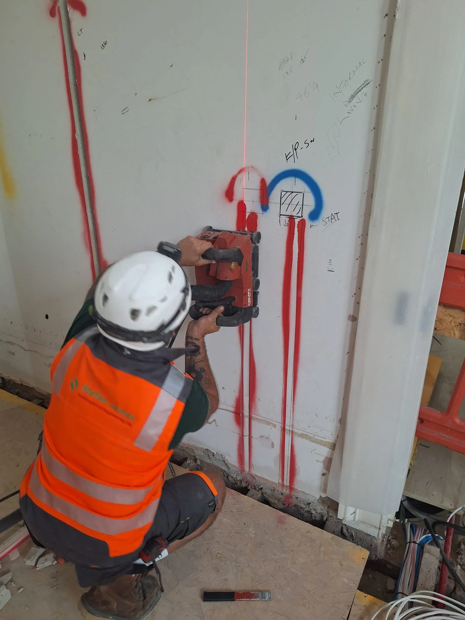 A construction worker in an orange safety vest and white helmet using a red power sander to work on a white wall with electrical wiring and markings. He is kneeling on a plywood surface, with a level tool on the floor nearby, and handwritten notes and lines are visible on the wall.