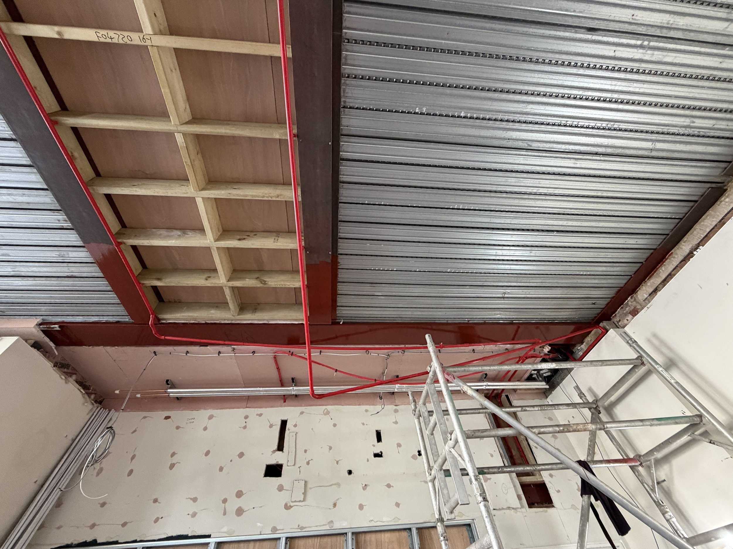 Construction site with exposed metal roof siding, wooden framing, and a drywall wall with hole cutouts, scaffolding, and electrical wiring visible.