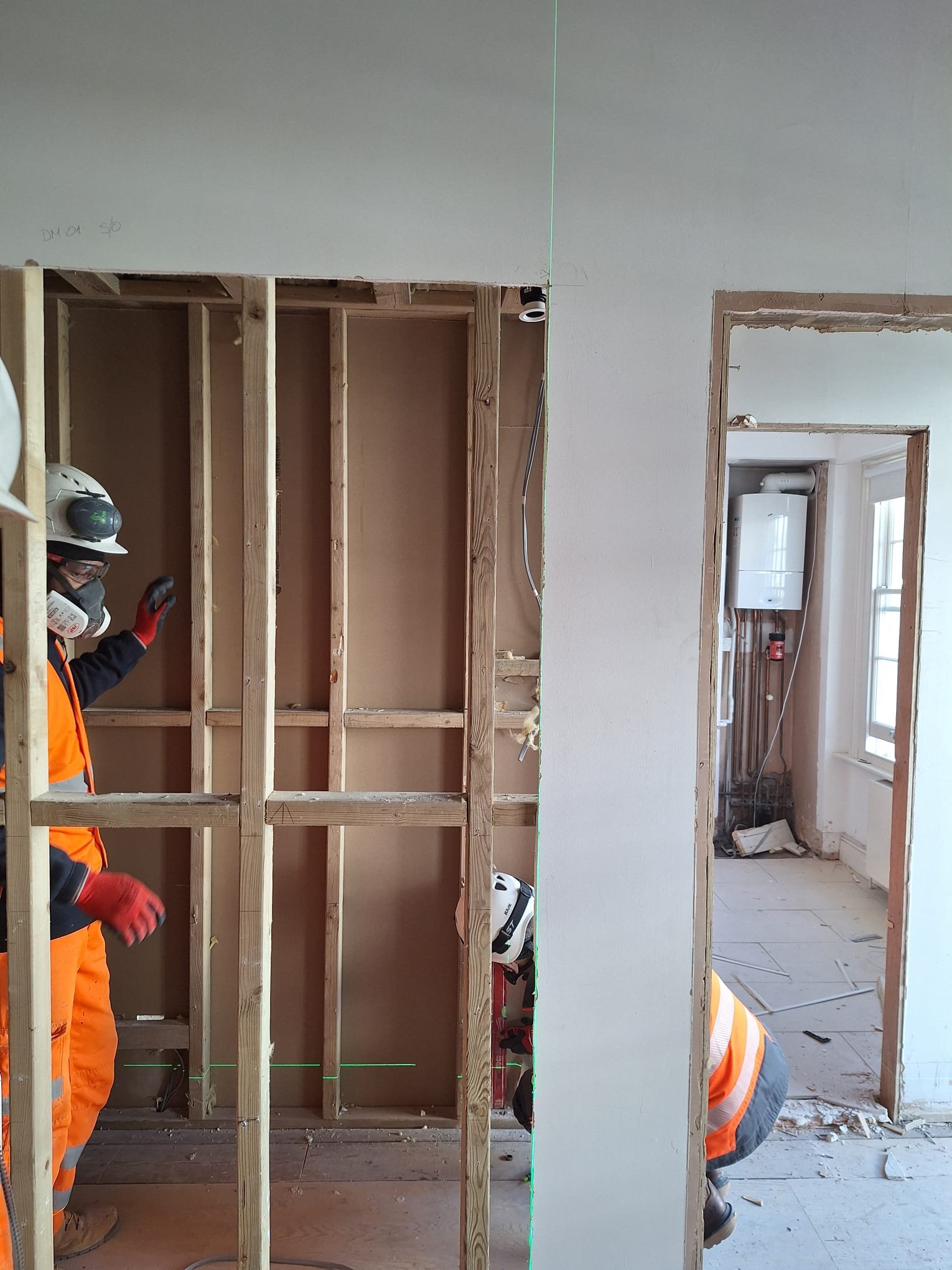 Construction workers wearing safety helmets and orange vests working on interior wall framing in a home renovation project. The photo shows wooden studs and partially finished drywall areas.