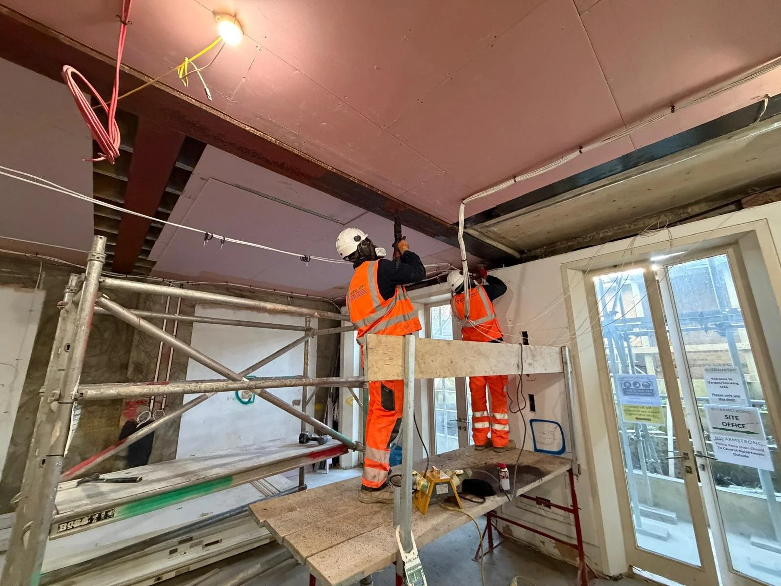 Two construction workers in orange safety vests and helmets working on installing or repairing electrical wiring near a ceiling inside a building. One worker is using a drill while the other is working nearby. There is construction scaffolding and tools around them, and a door with signs on it.