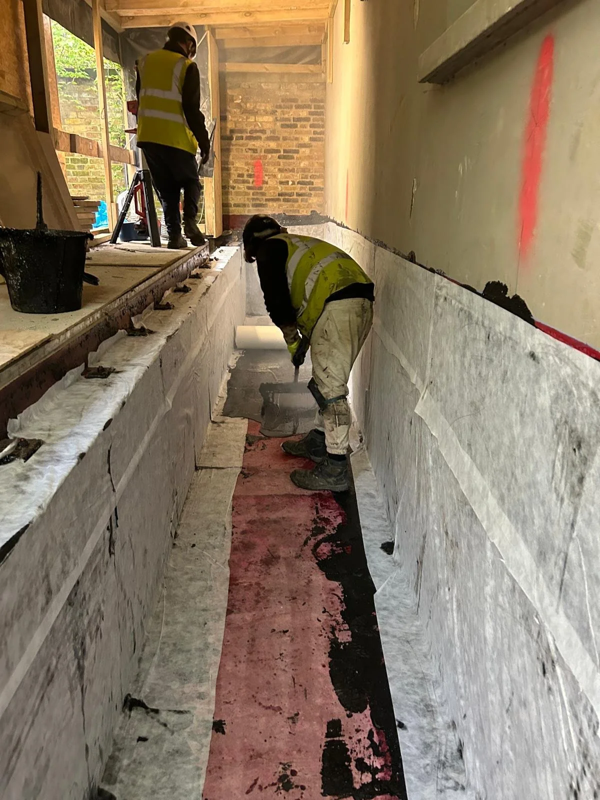 Two construction workers in safety vests working inside an unfinished building, installing or repairing a surface along a narrow, illuminated corridor lined with concrete or stone walls and an unfinished ceiling.