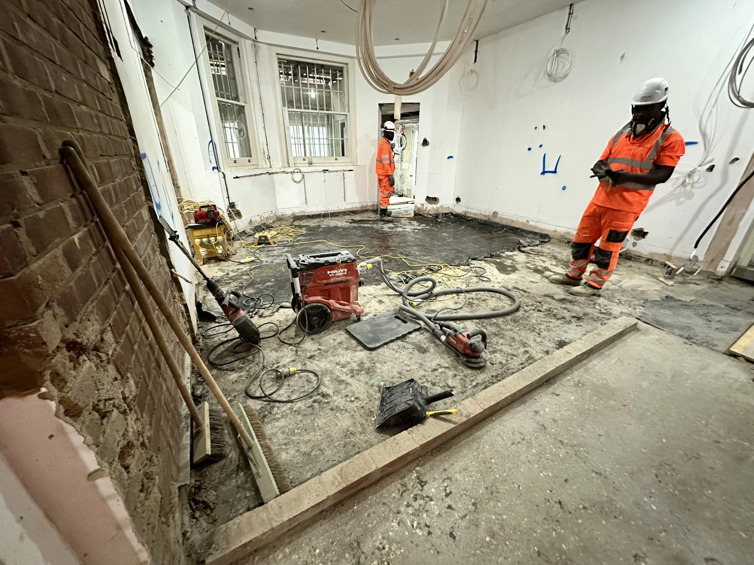 Two construction workers in bright orange safety gear inside a building where the flooring and walls are being renovated. The room has multiple large windows with bars, exposed wires hanging from the ceiling, and construction tools and equipment scattered on the floor.