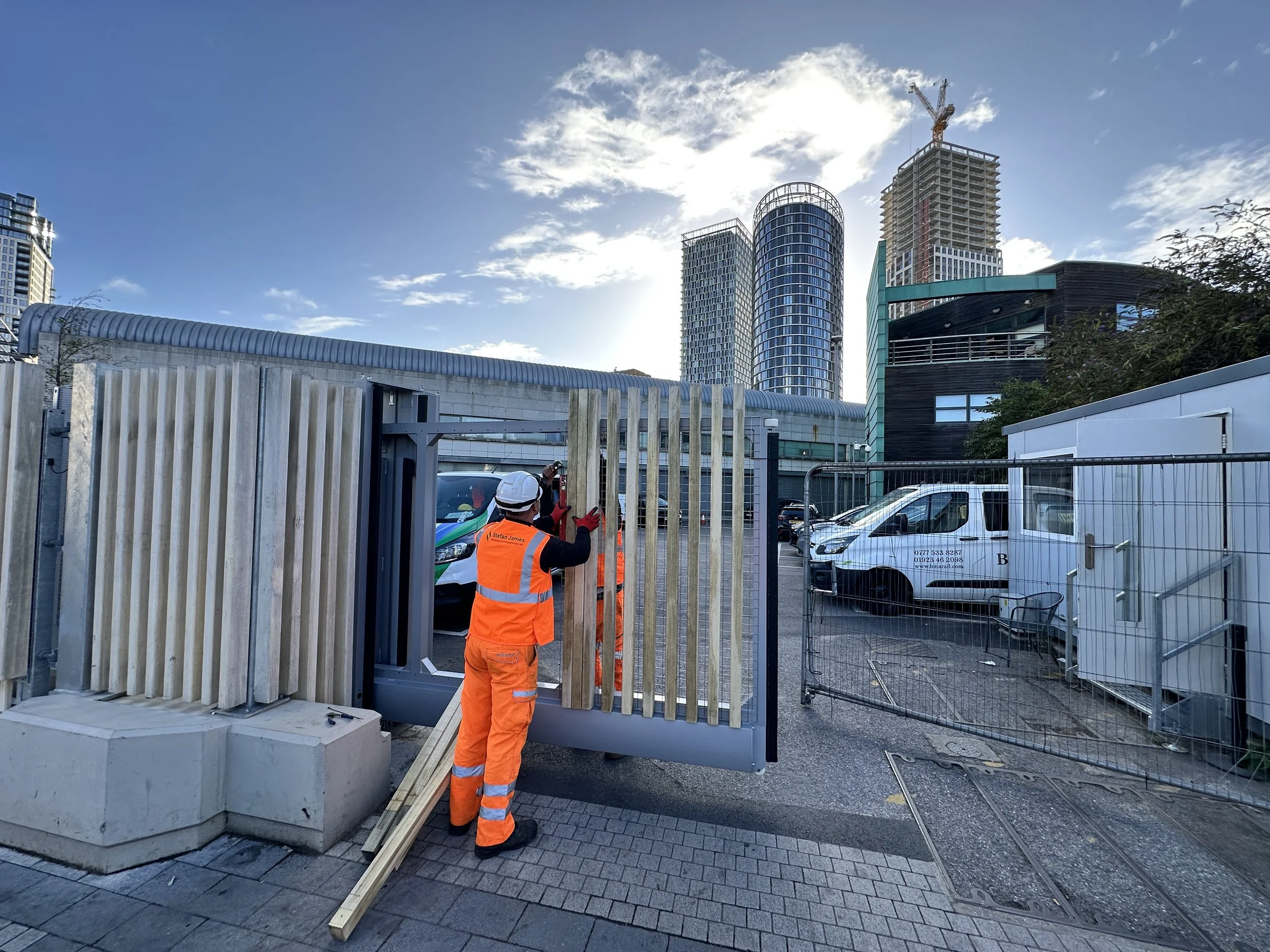 Construction worker in orange safety gear installing a fence panels in an urban area with high-rise buildings and a construction crane in the background.