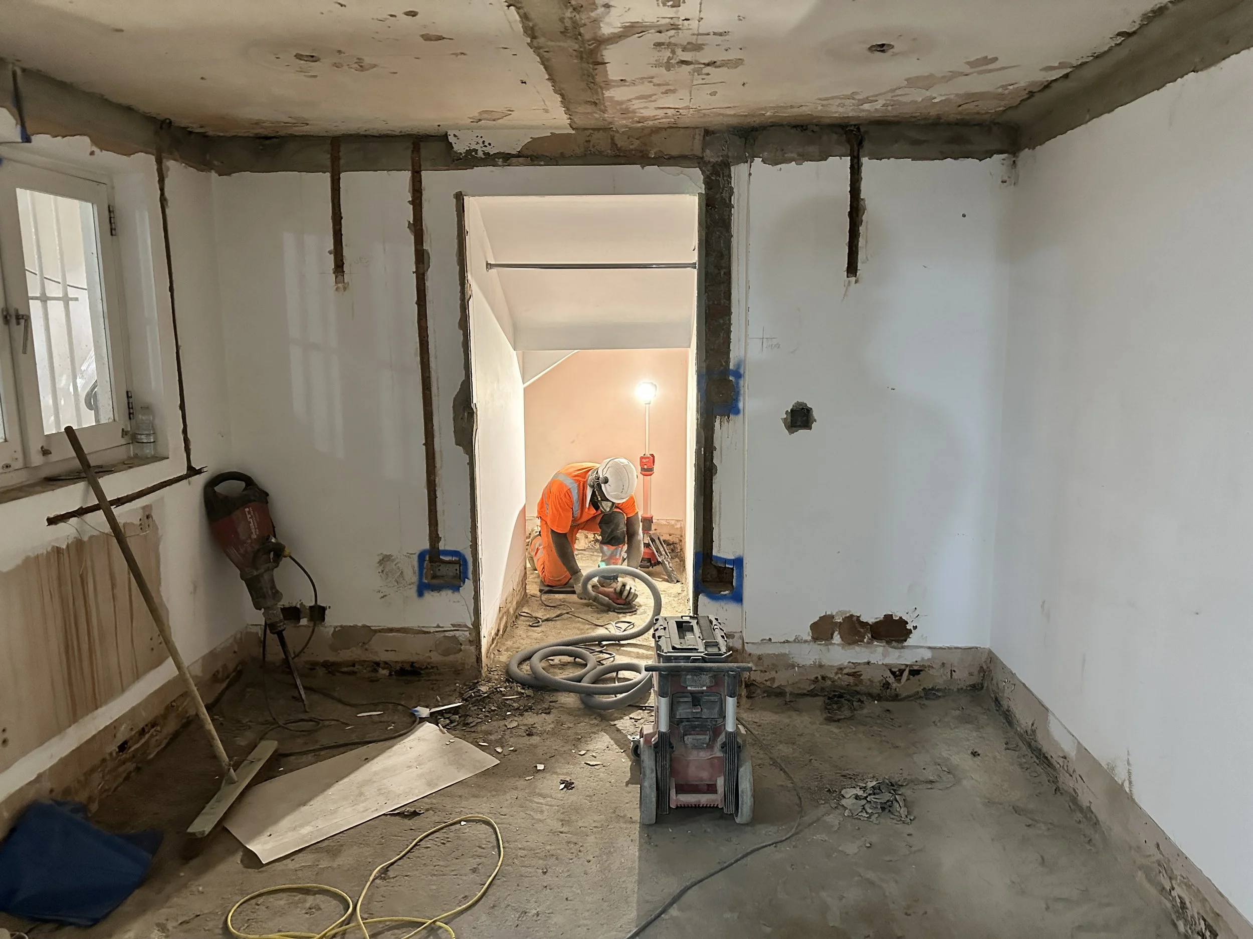 Construction worker wearing a helmet and orange safety vest working in a room under renovation. The room has exposed pipes, unfinished walls, and construction tools on the floor.