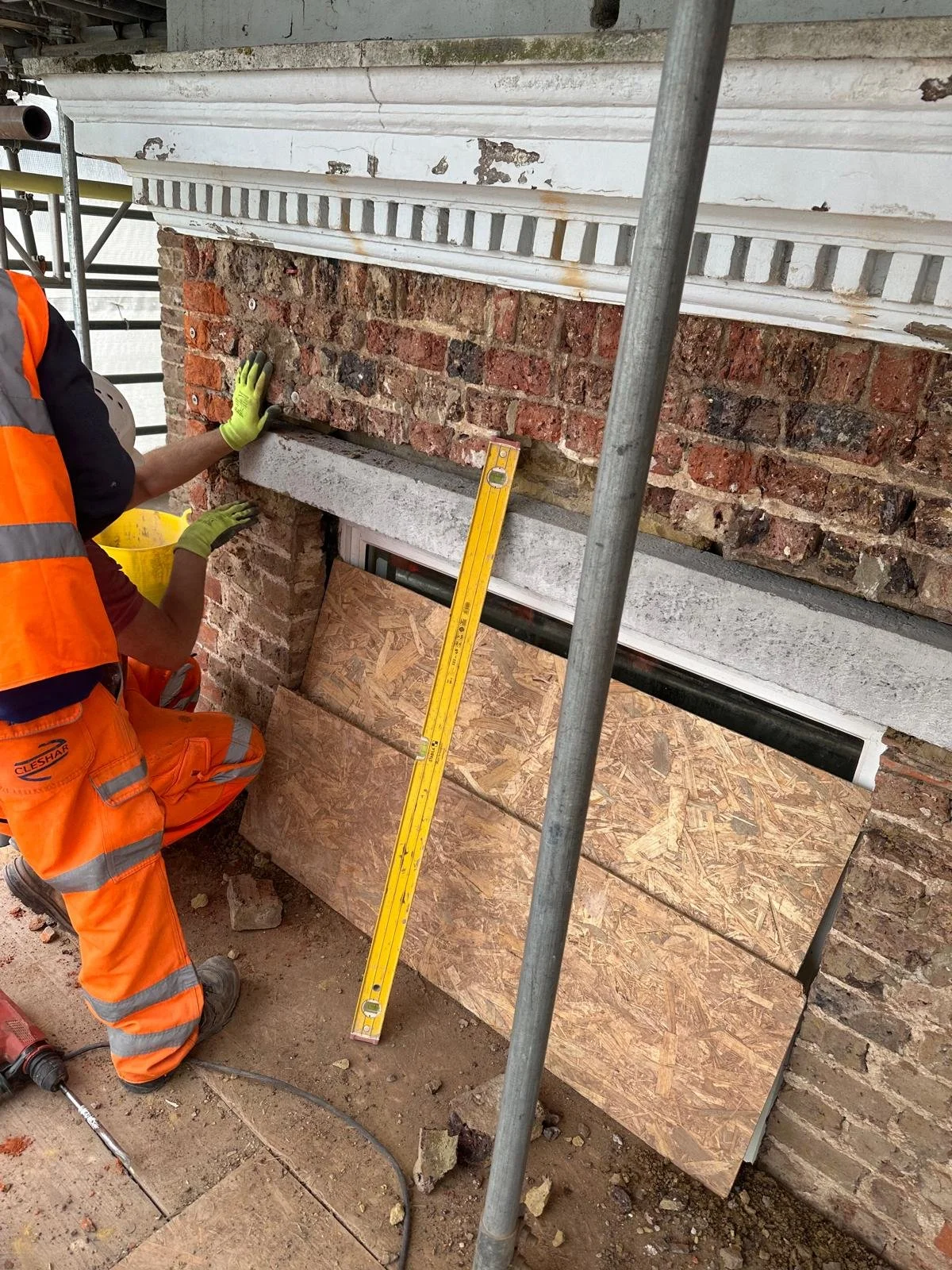 Construction workers installing a new brick wall, with a yellow level tool leaning against the wall, and a piece of plywood at the base. One worker is kneeling and holding the wall, wearing an orange safety vest and gloves.