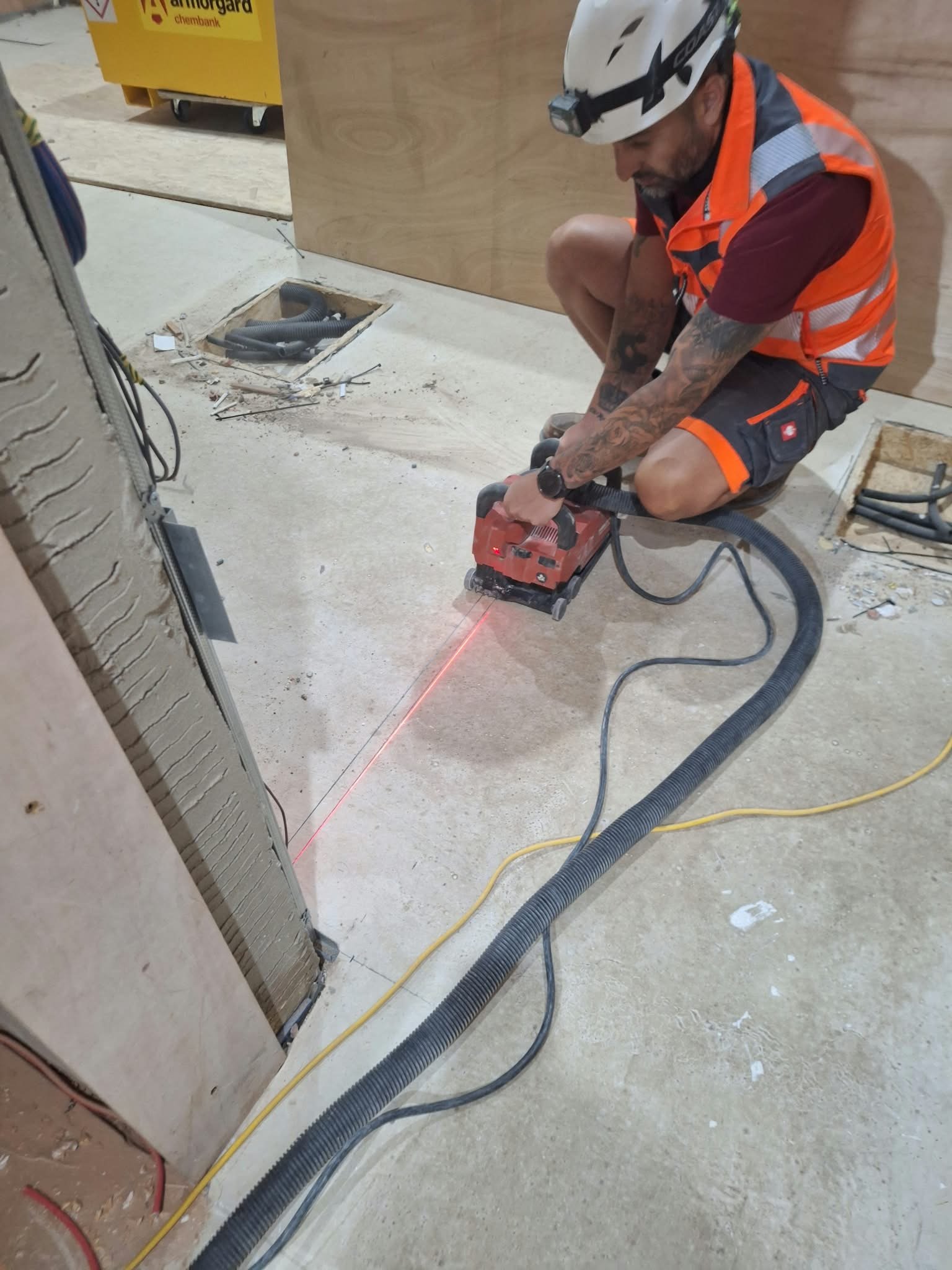A construction worker wearing a helmet with a headlamp, orange safety vest, and dark shorts, crouching and using a tool to cut a line of fiber optic cables on a concrete floor. There are construction materials and an open electrical box nearby.