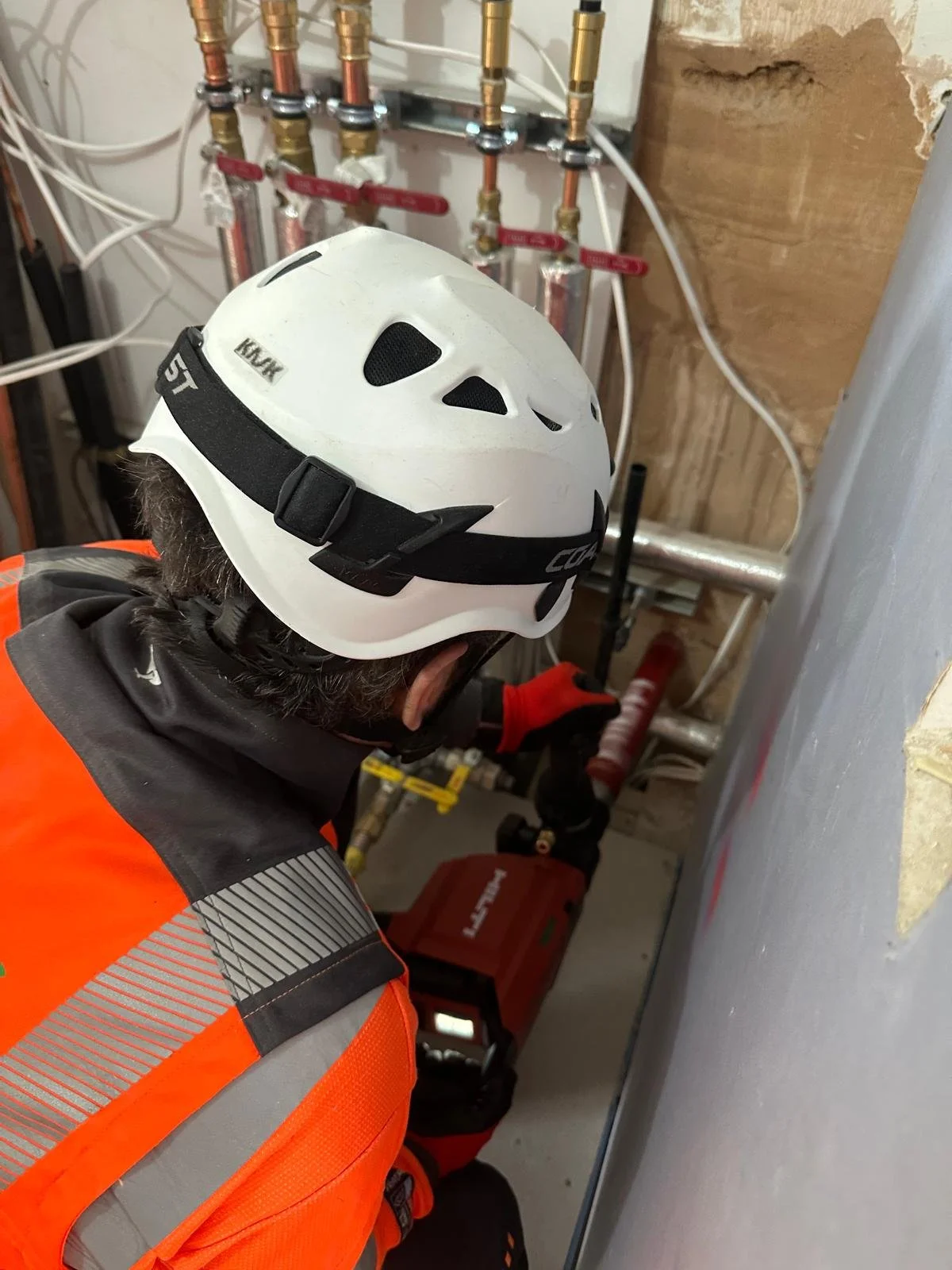A worker wearing an orange safety vest, black jacket, and white helmet inspecting plumbing pipes in a utility room.